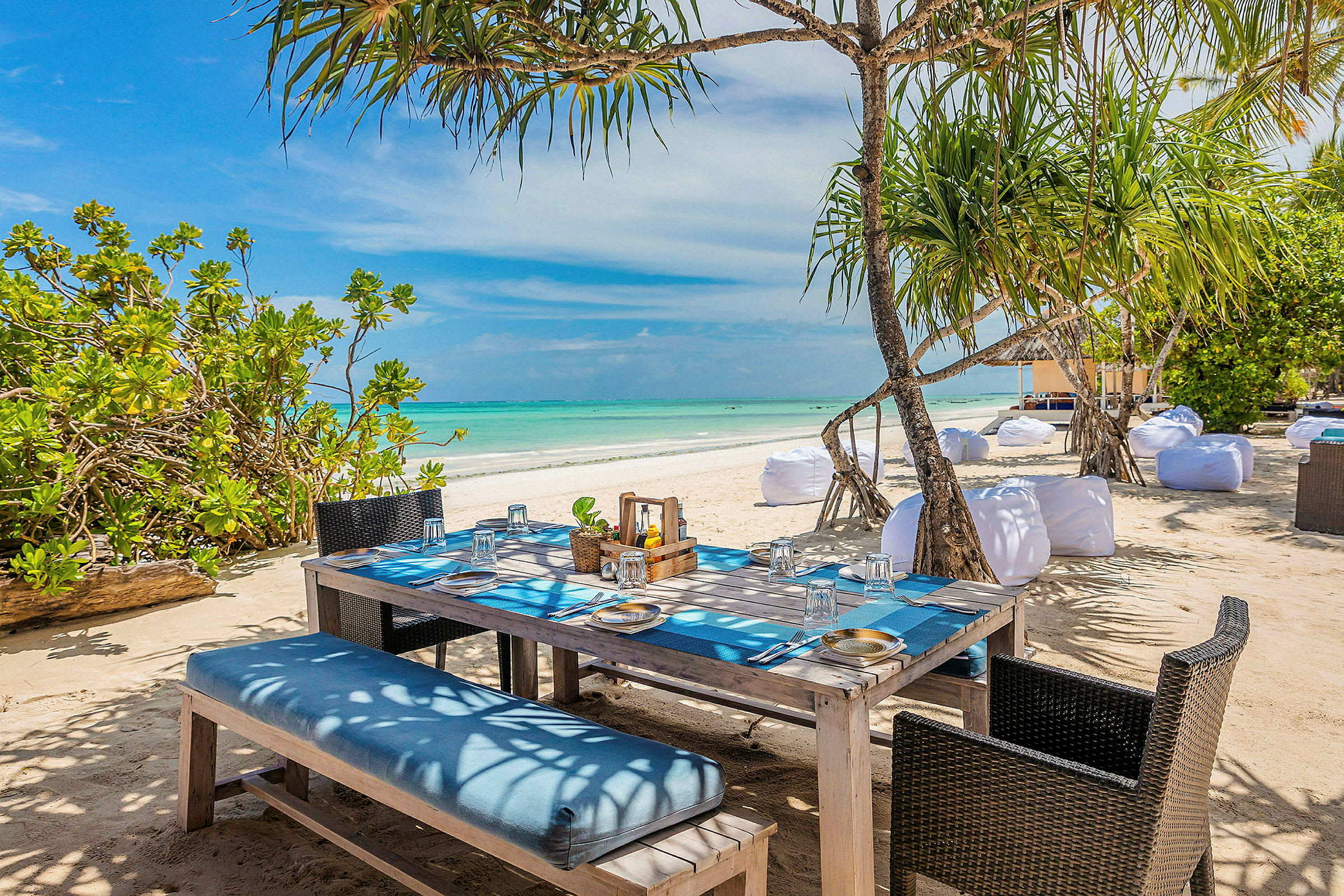 Ett uppdukat bord med duk och glas ståendes på en sandstrand med närliggande träd och utsikt över det turkosa havet. (AI-genererad text)