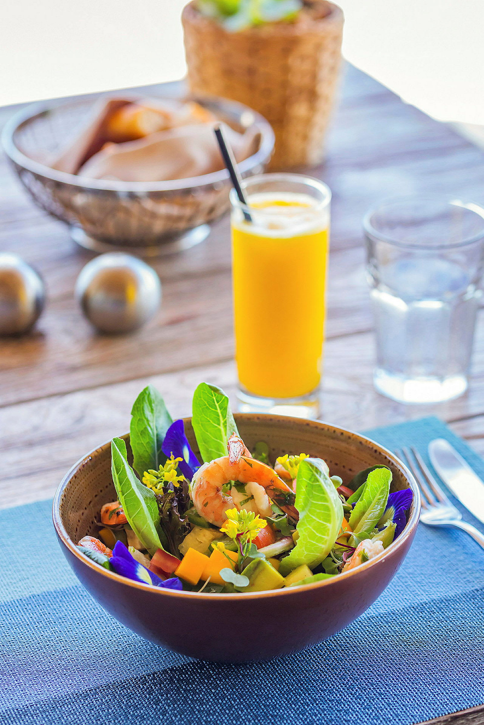 En sallad i en skål med räkor, gröna blad och blommor. Ett glas apelsinjuice och bröd i bakgrunden. (AI-genererad text)