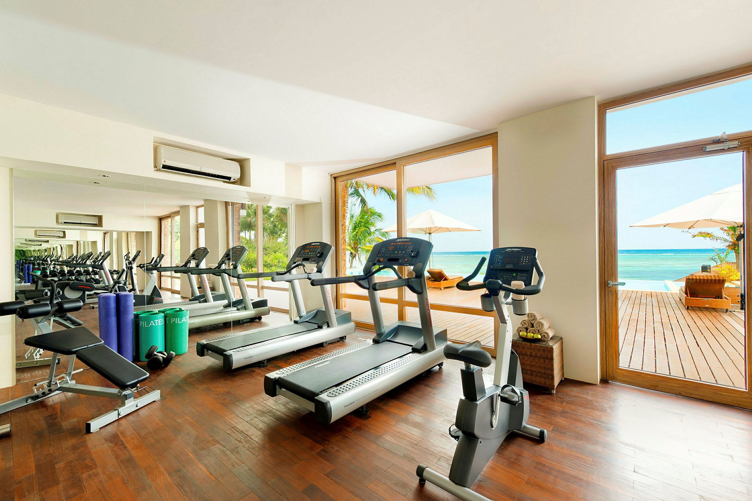 Ett gym med träningsmaskiner och spegelvägg, stora fönster öppnar upp mot en terrass med utsikt över havet. (AI-genererad text)