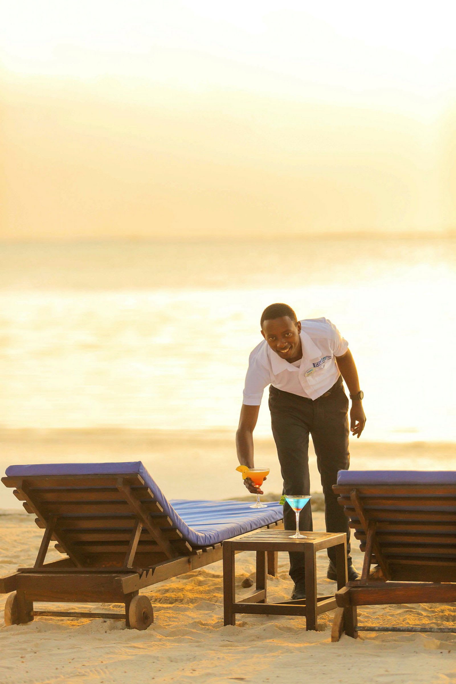 En person placerar en cocktailglas på ett bord mellan två solstolar på stranden. (AI-genererad text)