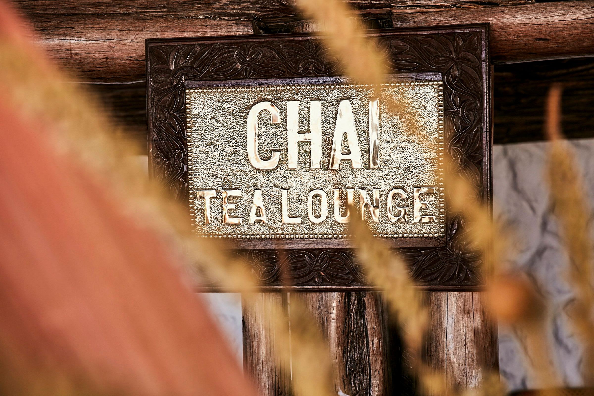 En närbild av en skylt som det står 'Chai Tea Lounge' på, omgiven av trädekorationer och vissnande gräs i förgrunden. (AI-genererad text)