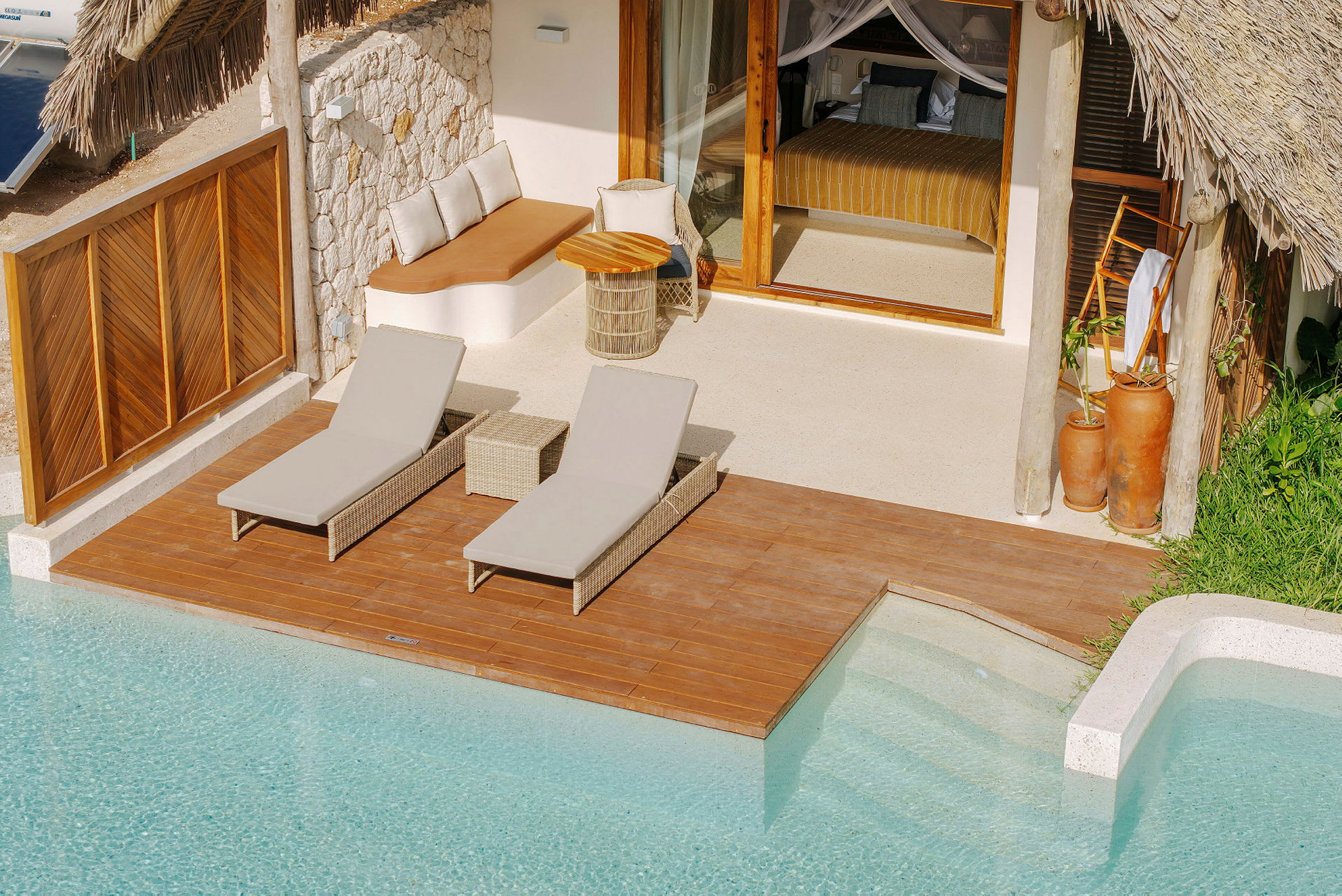Ett trädäck vid en pool med två solstolar och en loungeyta. Bakom däckområdet finns en villa med glasdörrar och halmtak. (AI-genererad text)