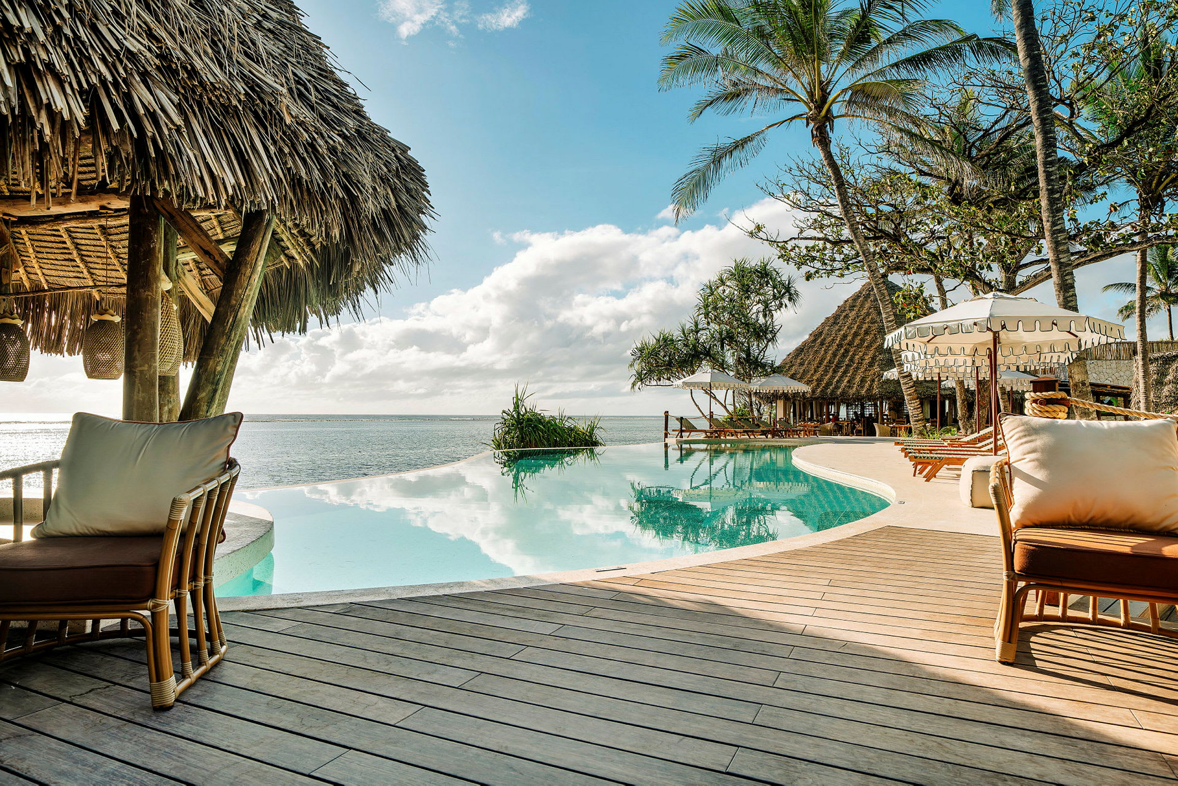 Bild av en infinitypool intill en strand med halmtak, solstolar och parasoller i en tropisk miljö. (AI-genererad text)