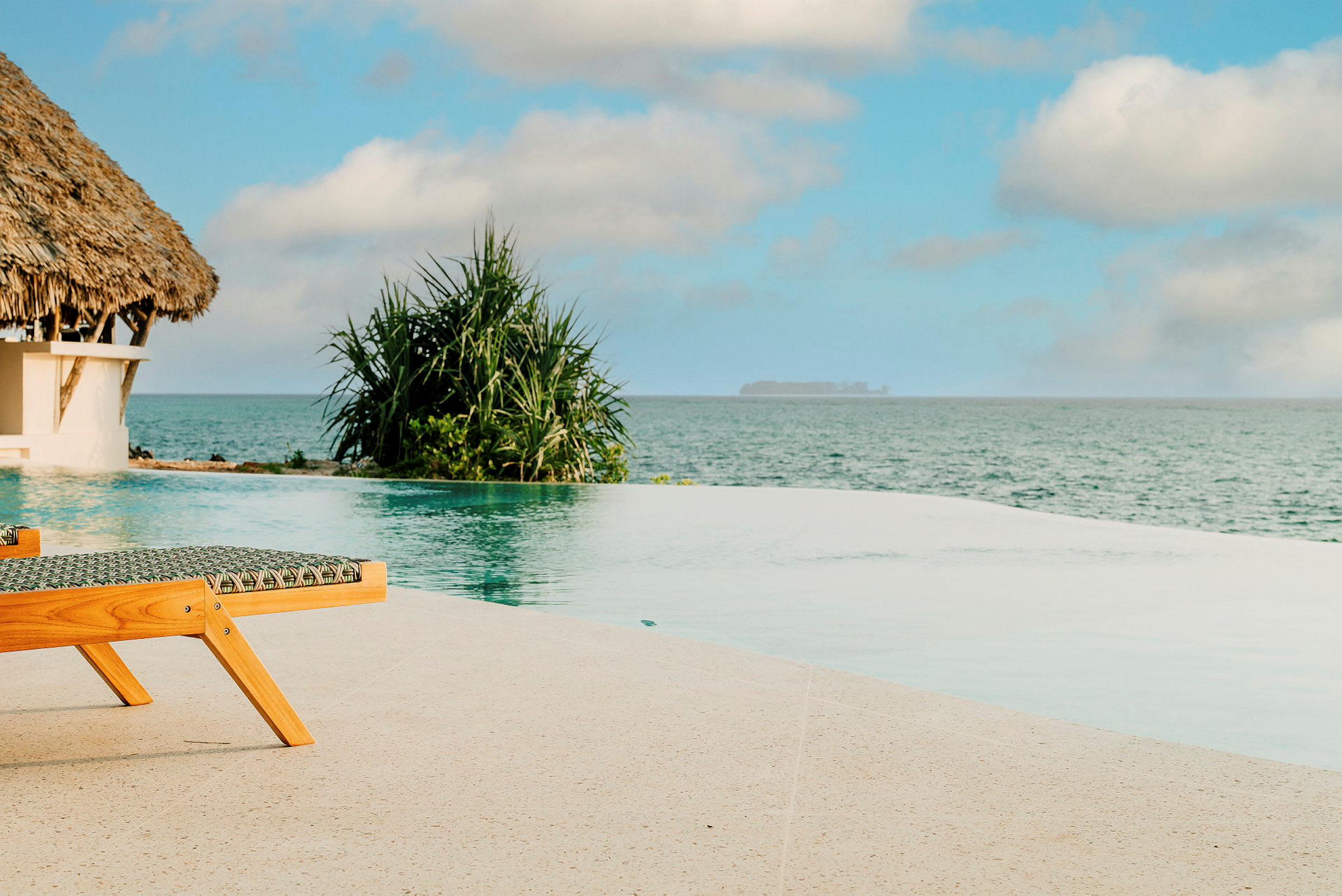En infinitypool vid havet, med en solstol och ett hus med halmtak i bakgrunden. (AI-genererad text)