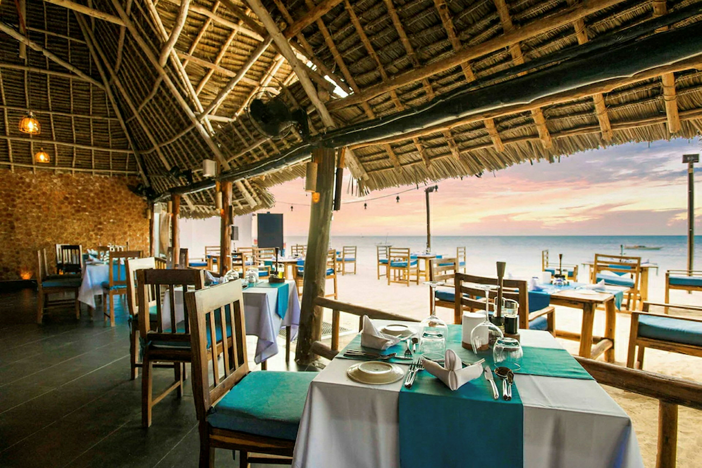 Bild av en strandnära restaurang med bord och stolar under ett halmtak. Havet och solnedgången syns i bakgrunden. (AI-genererad text)