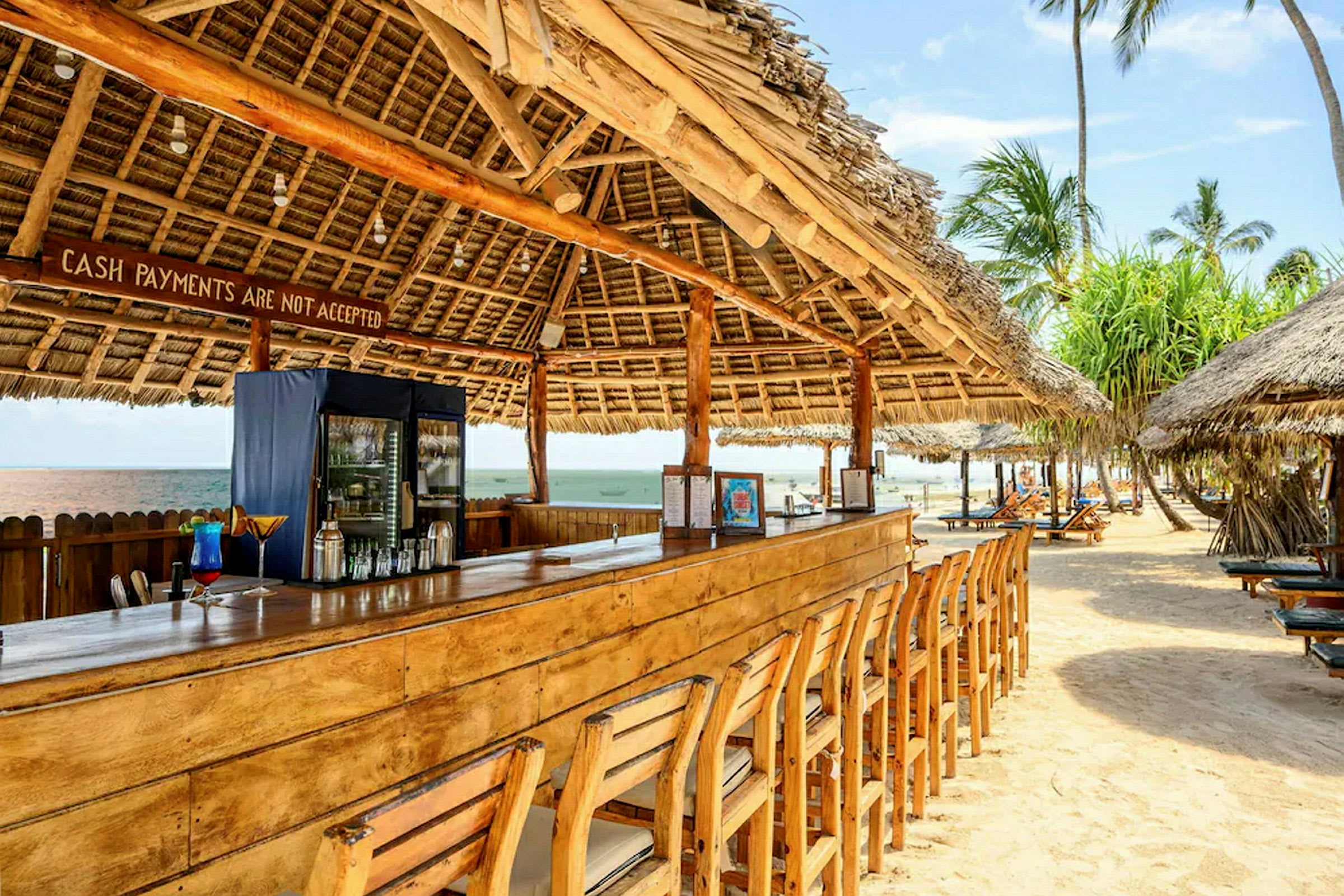 Strandbar med träbord och stolar under tak av palmblad. (AI-genererad text)