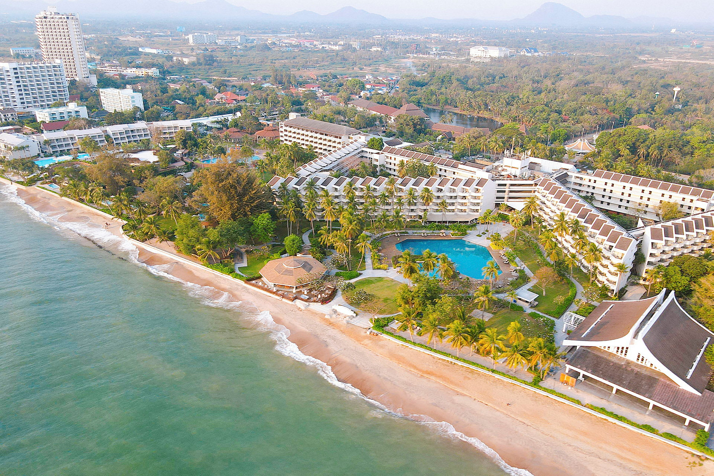 Flygfoto av en resort vid stranden med byggnader, palmer och pooler. (AI-genererad text)
