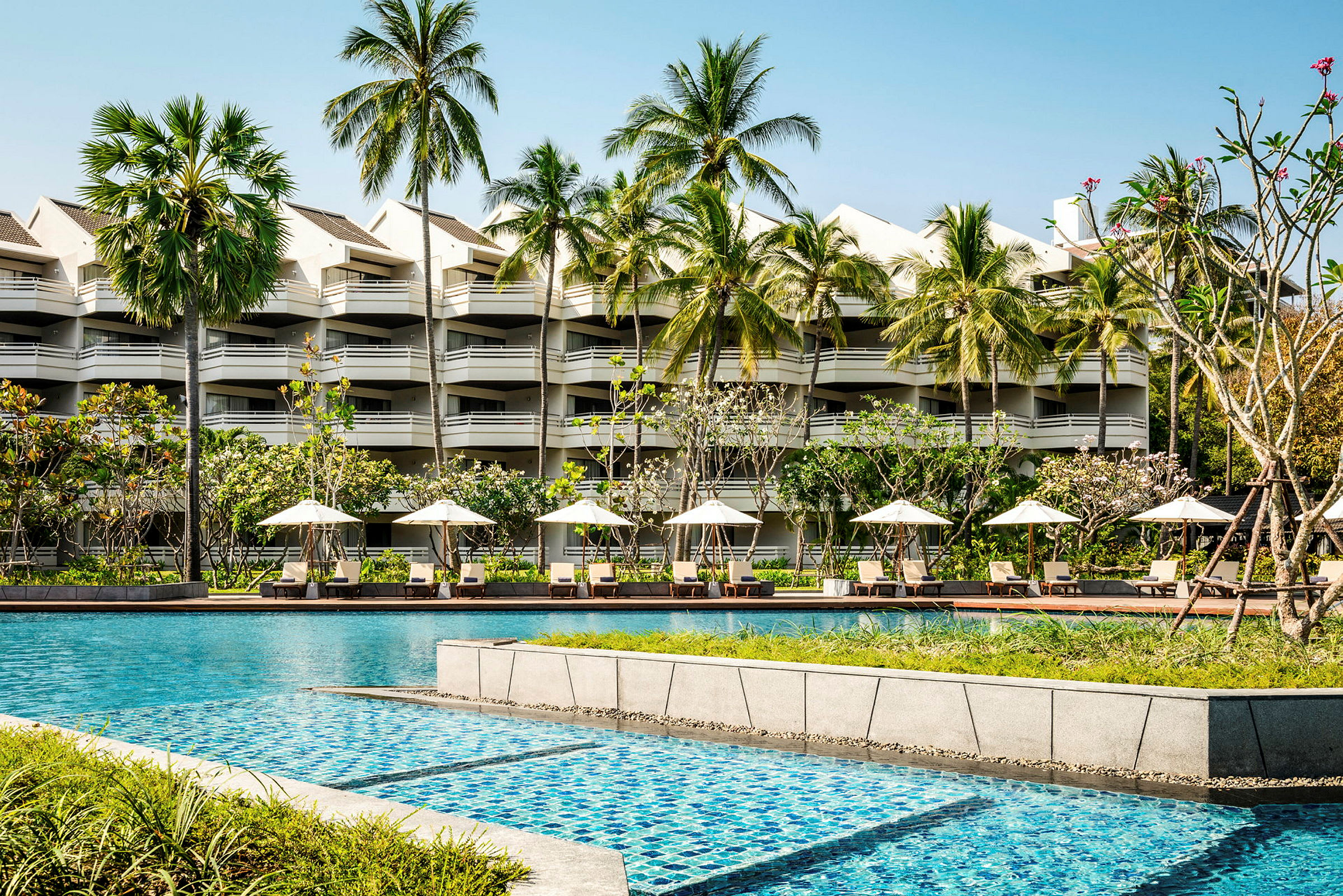 Hotellbyggnad med flera våningar bakom en pool och solstolar med parasoller, omgiven av palmer och grönska. (AI-genererad text)