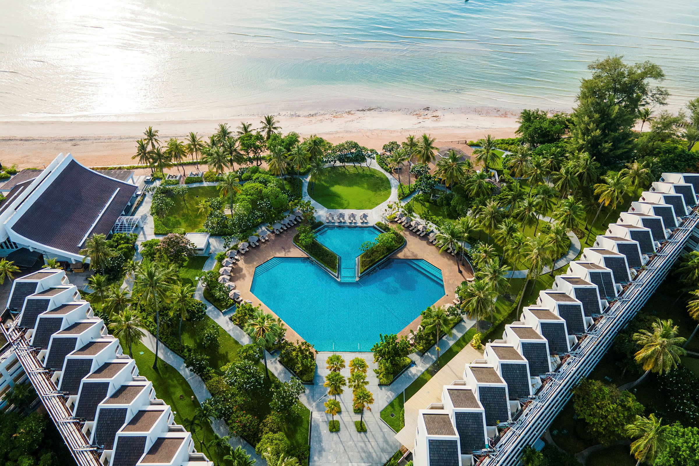 Flygvy över en resort med swimmingpool, grönska, och en strand i bakgrunden. (AI-genererad text)