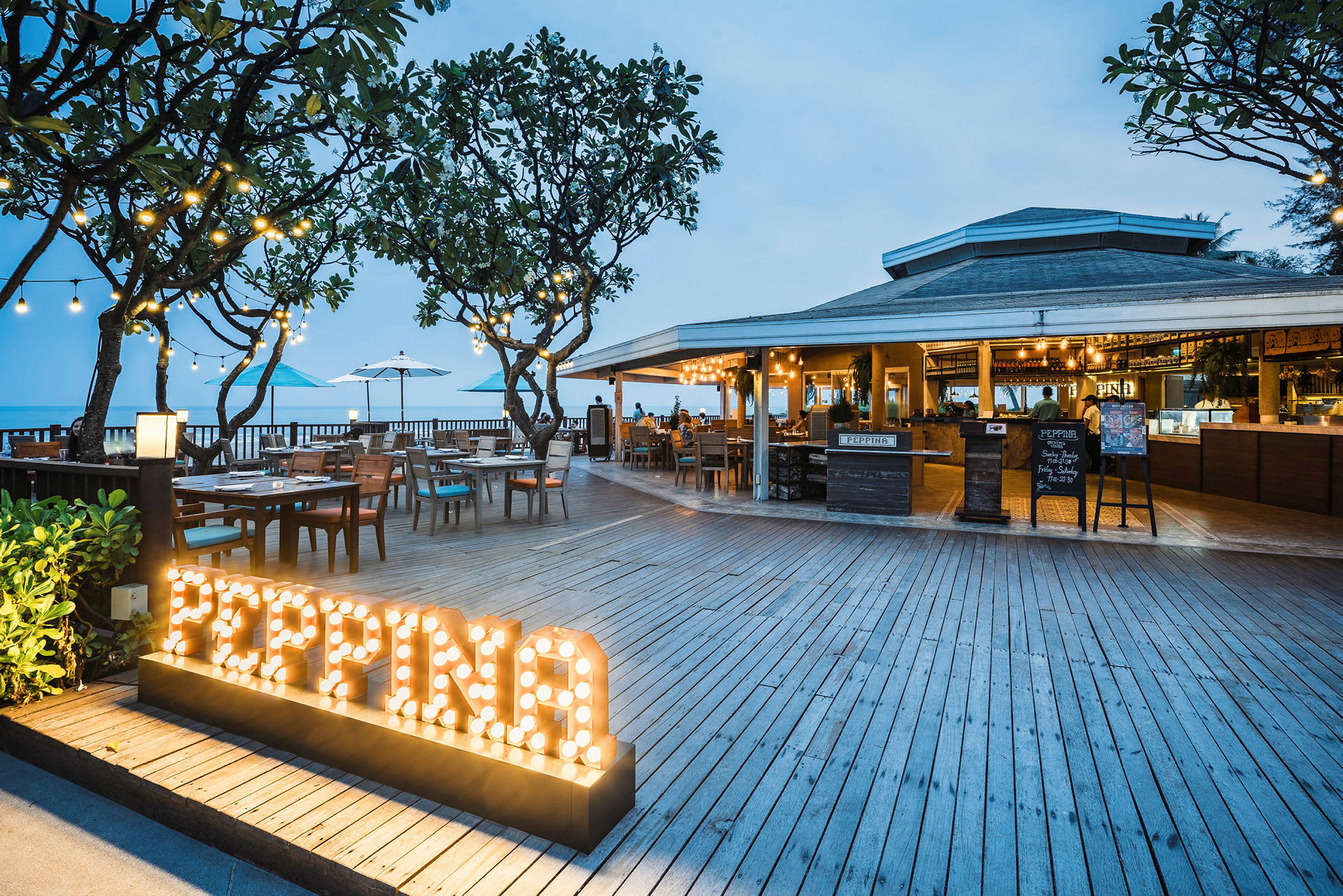 En restaurang med trädäck, bord, och sittplatser utomhus nära havet. Skylt med upplyst texten 'PEPPINA'. (AI-genererad text)