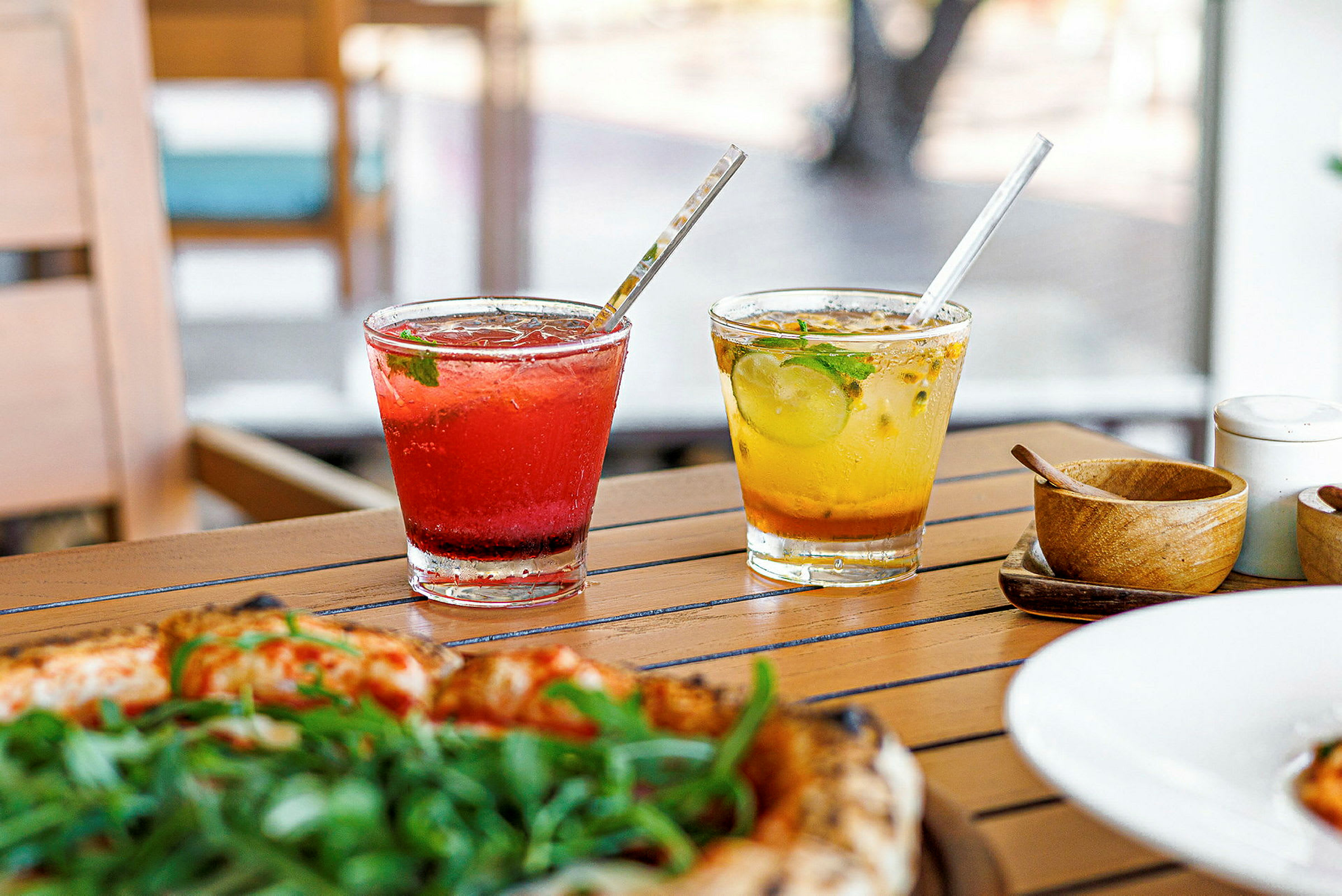 Två glas med färgglada drycker står på ett träbord. Ett glas innehåller röd dryck och det andra glaset gul dryck. Framför glasen syns en pizza med grön topping. (AI-genererad text)