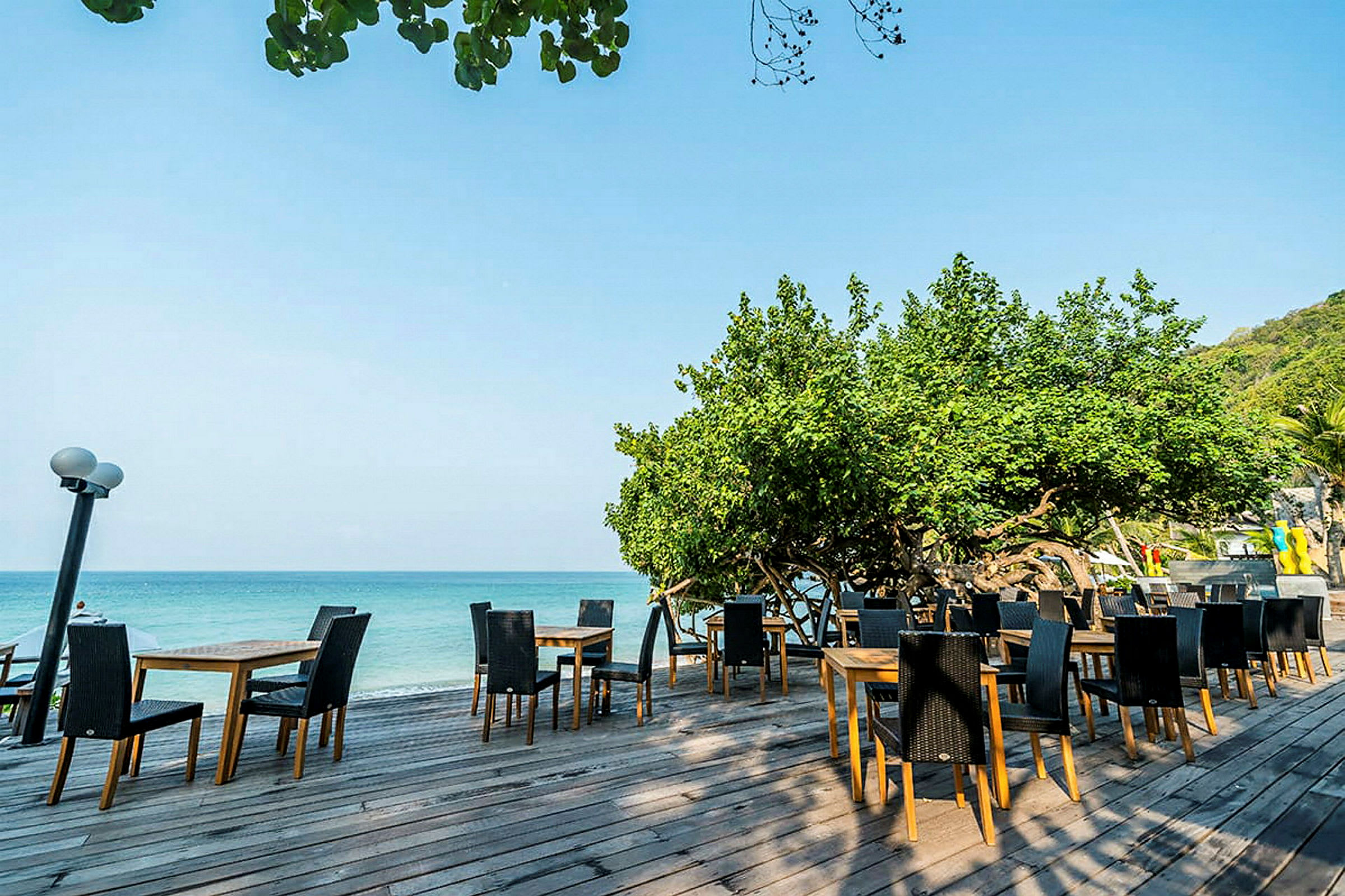 En utomhusrestaurang med bord och stolar placerade på en träaltan vid havet, omgiven av grönska. (AI-genererad text)