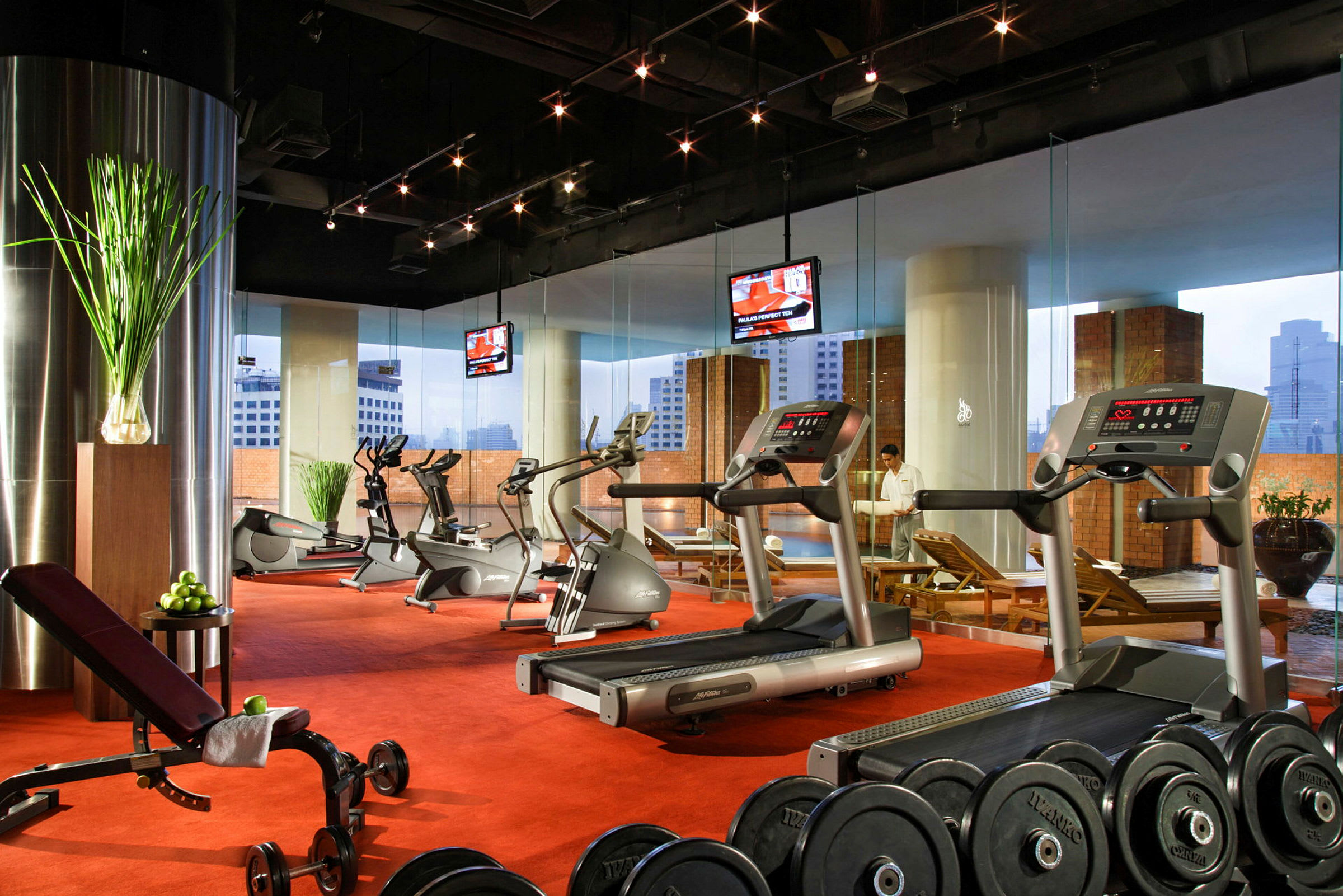 Ett gym med träningsmaskiner, fria vikter, och motionscyklar, med stora fönster och utsikt över stadens skyline. (AI-genererad text)