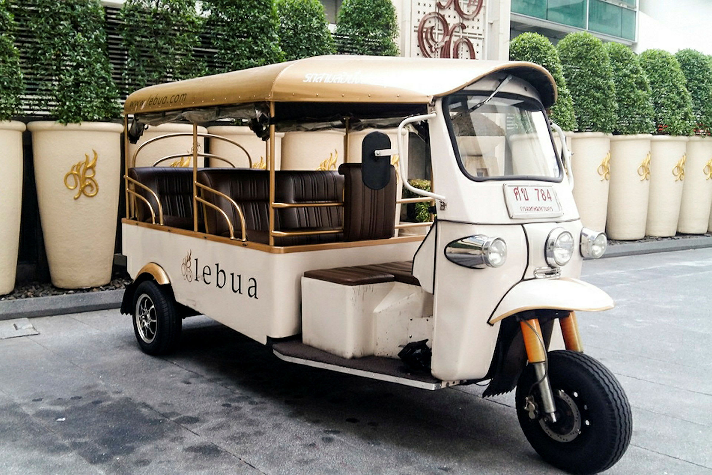 En vit tuk-tuk med beige tak står parkerad bredvid en rad stora vita krukor och gröna växter i bakgrunden. (AI-genererad text)