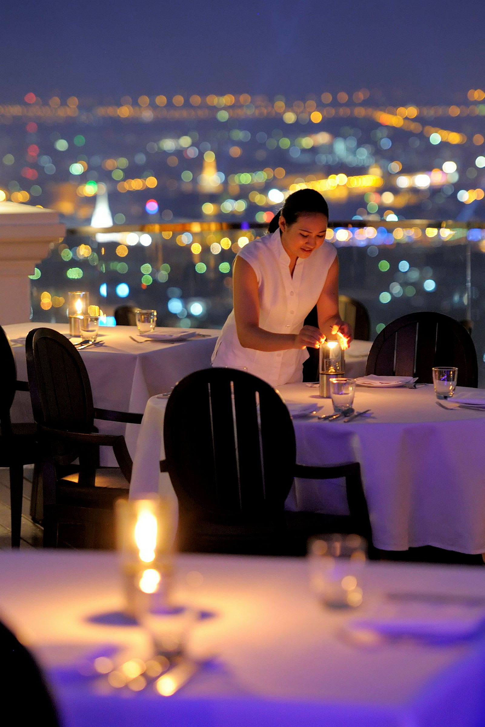 En person tänder ljus på ett dukat bord i en restaurang med utsikt över en stads silhuett. (AI-genererad text)