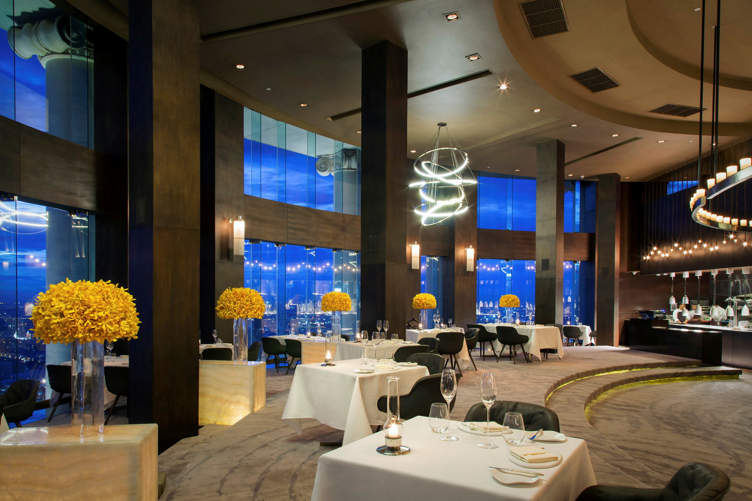 En elegant restaurang med dukade bord, moderna möbler och stora fönster som vetter mot en stad. Gula blomdekorationer är placerade på piedestaler. (AI-genererad text)