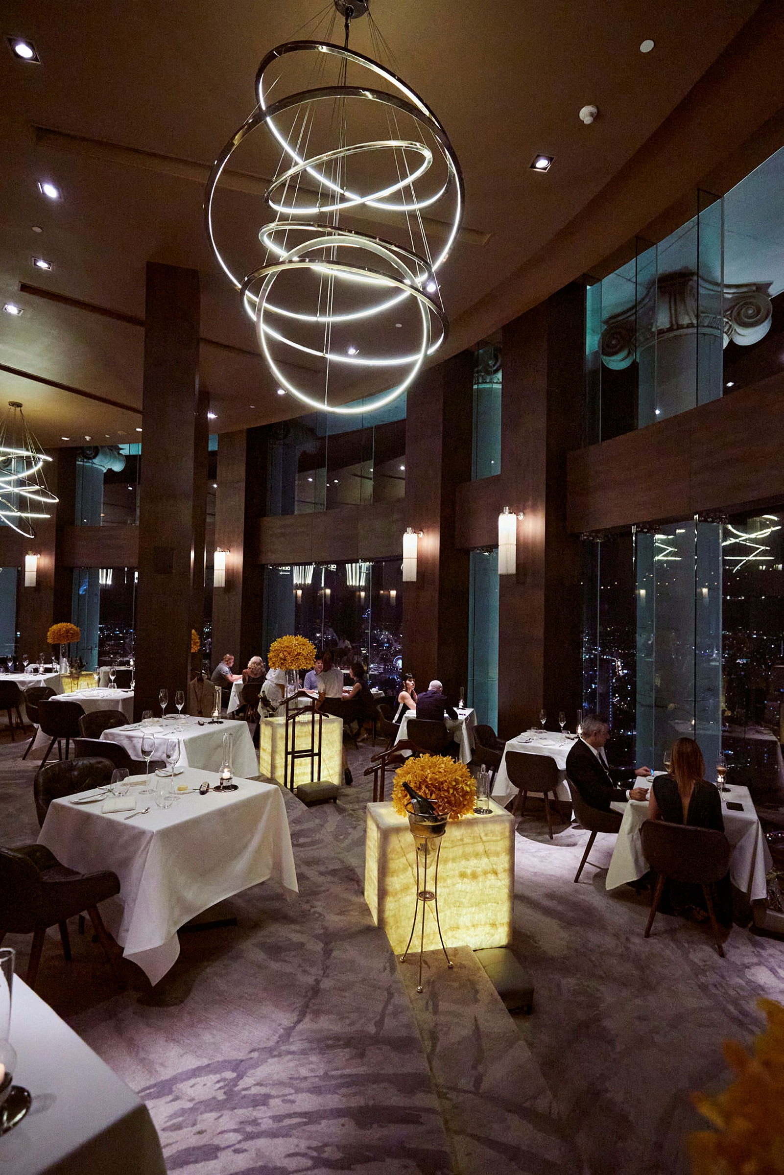 En lyxig restaurang med fönster som visar stadsvyer på kvällen. I förgrunden syns bord med bordsdekorationer. (AI-genererad text)