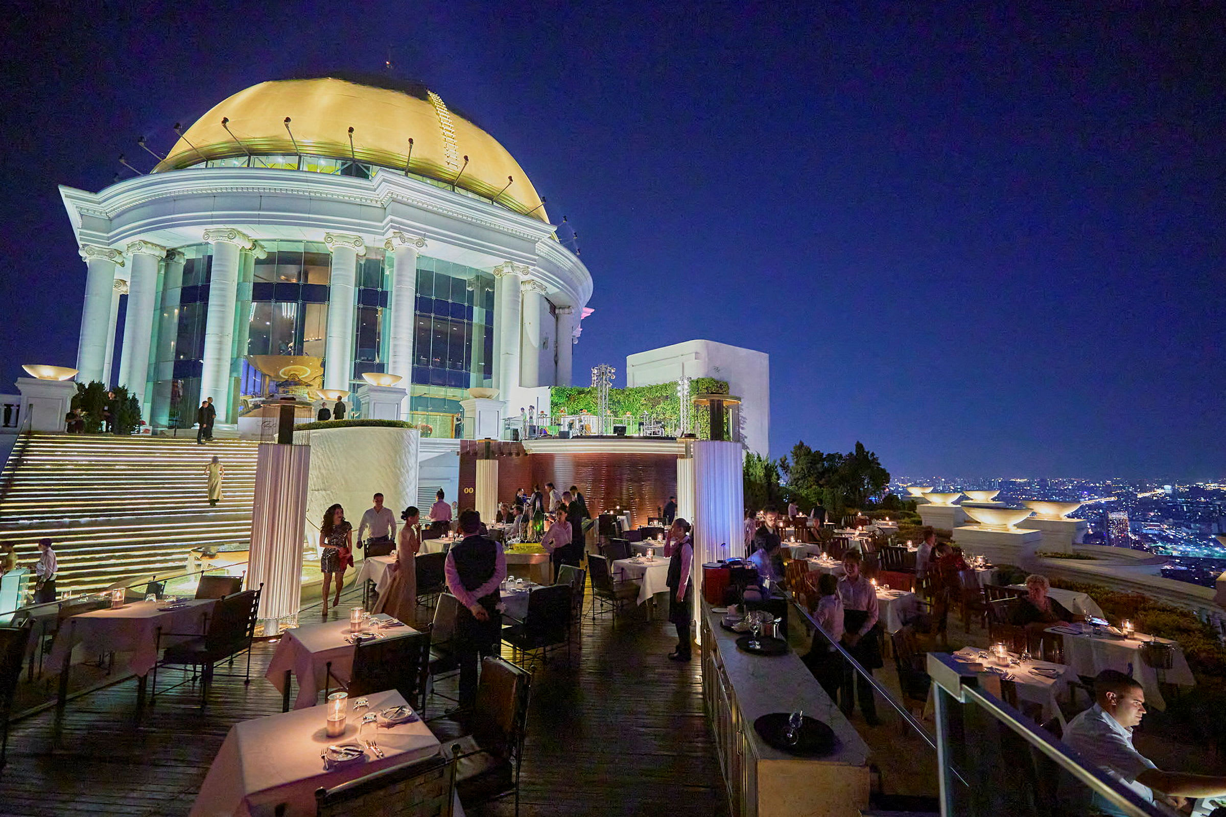 En restaurang vid en takterrass med utsikt över en upplyst stad i skymningen. På bilden syns bord, gäster och servitörer. (AI-genererad text)