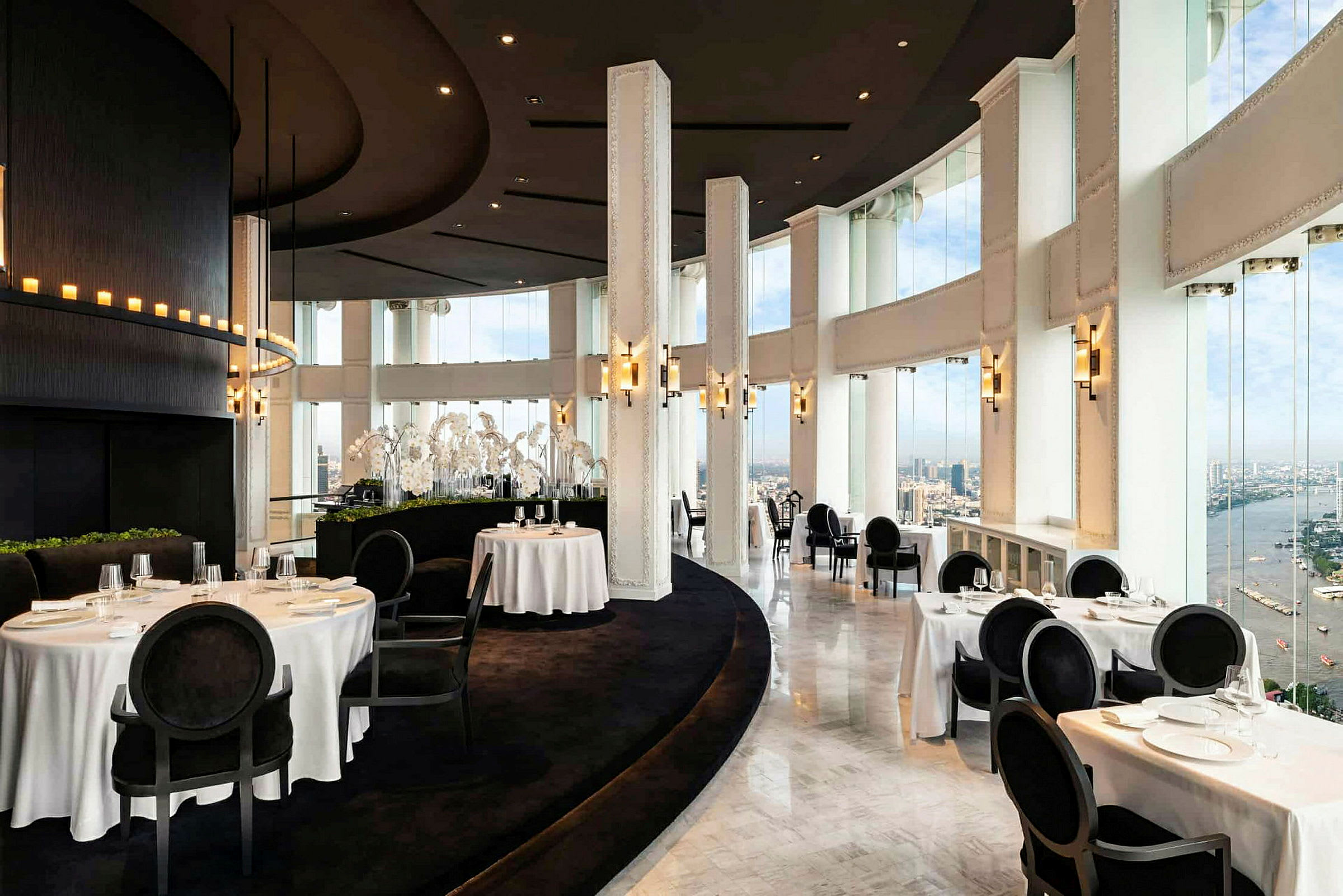 En elegant restaurang med höga fönster och vit dukning, som erbjuder utsikt över staden och en flod. (AI-genererad text)