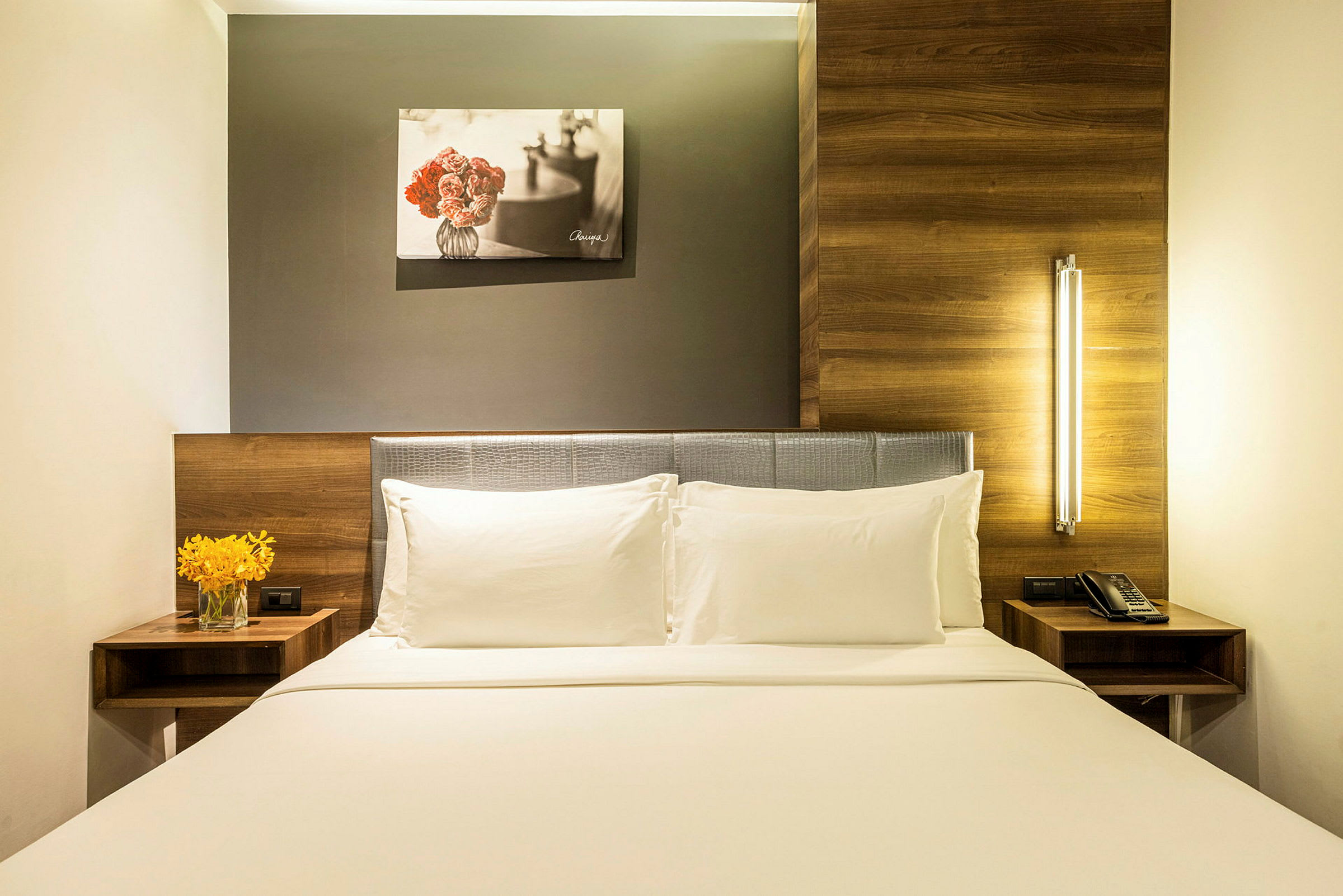 Ett hotellrum med en bekväm dubbelsäng, två vita kuddar och en vit sängklädsel. Ett nattygsbord med en vas med gula blommor till vänster och ett annat nattygsbord med en telefon till höger. Ovanför sängen hänger en målning av blommor. (AI-genererad text)