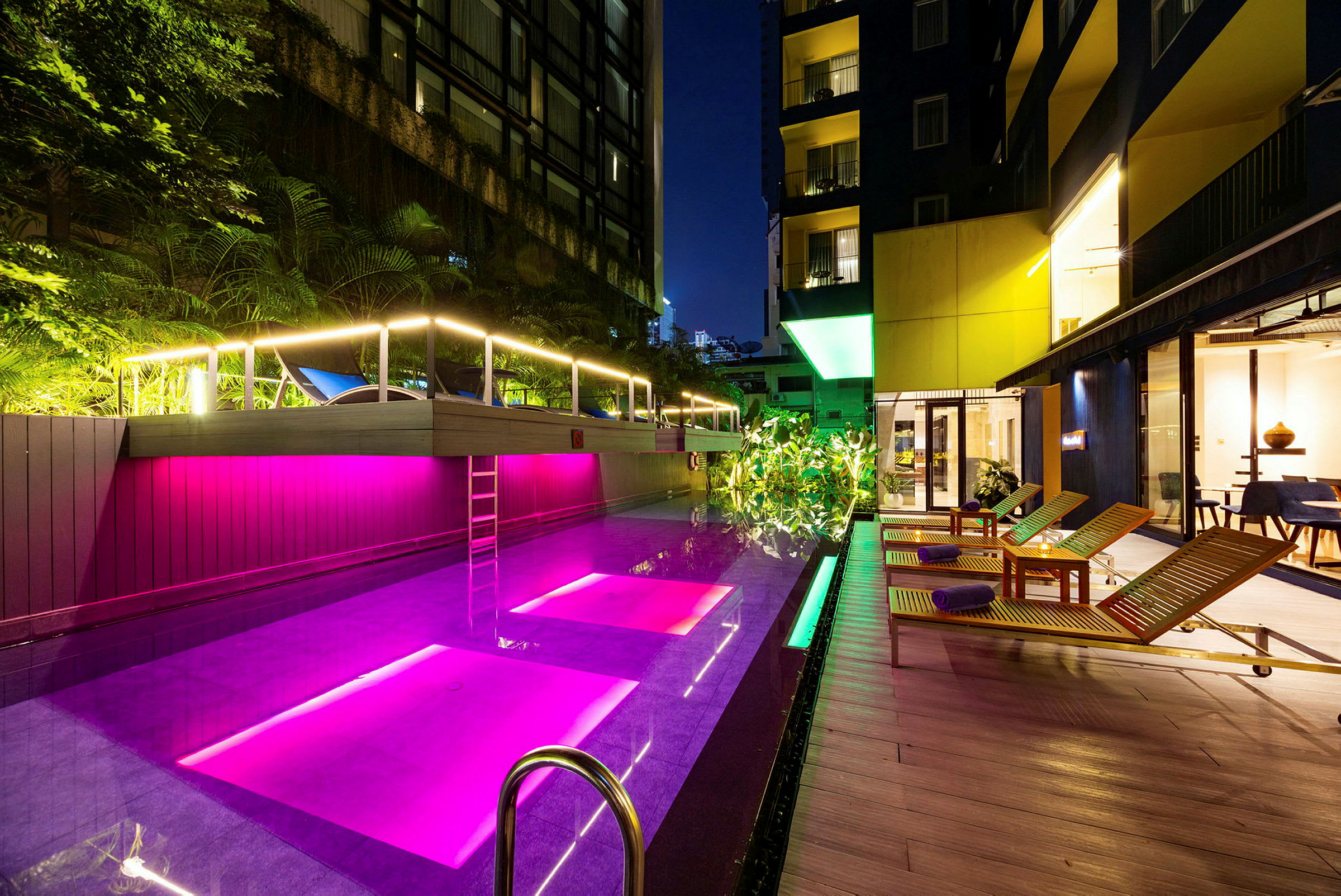 En pool med färgglada ljus och solstolar bredvid, omgiven av modern arkitektur och träd. (AI-genererad text)