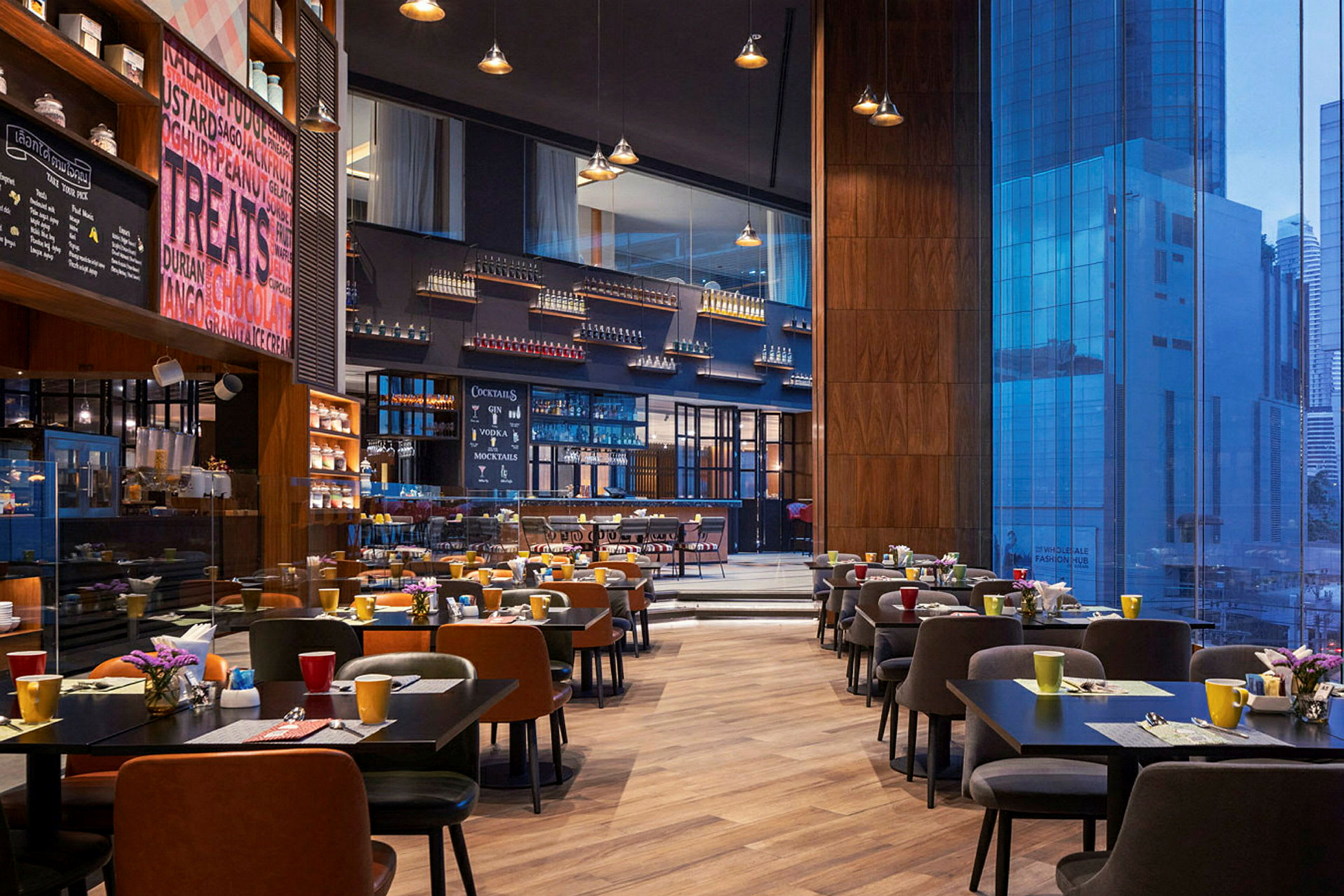 En rymlig restaurang med moderna bord och stolar uppställda, färgglada koppar och blommor på borden och en stor glasvägg som visar utsikt över stadens byggnader. (AI-genererad text)