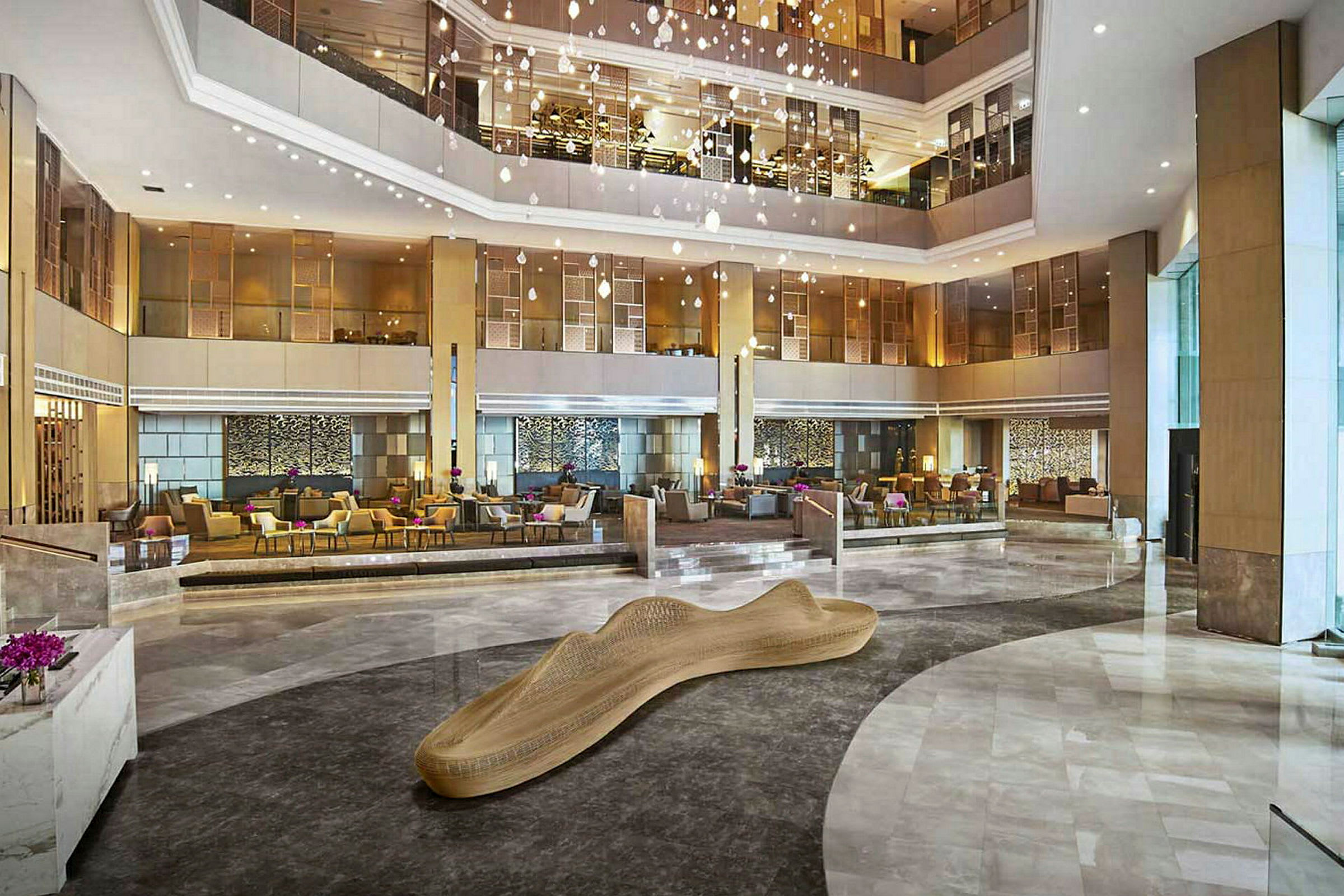 Ett stort hotellobbyområde med moderna möbler och en dekorativ träskulptur i mitten. Flera sittgrupper och elegant belysning. (AI-genererad text)