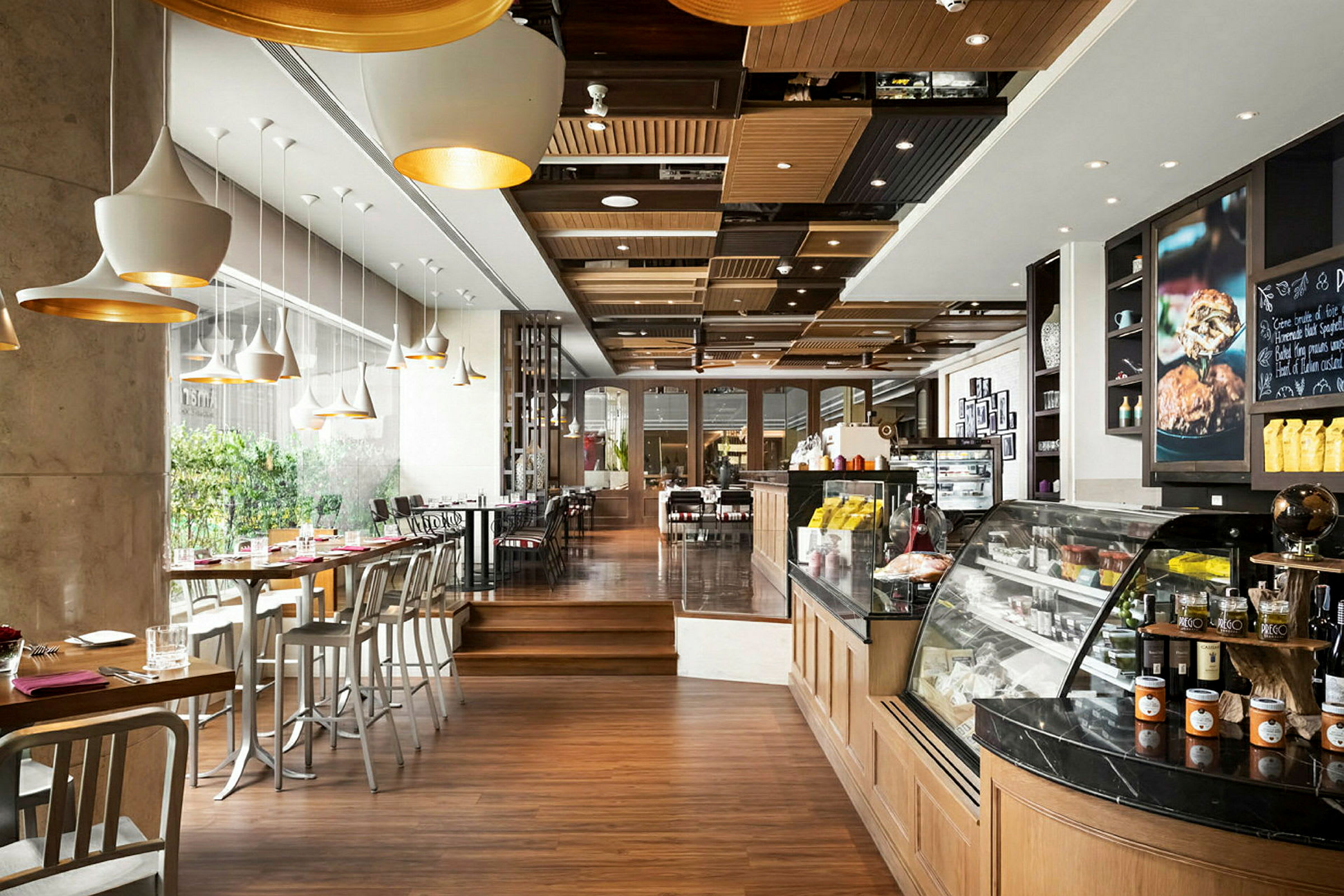 Ett modernt kafé med trägolv, stora fönster och sittplatser vid bord och barstolar. En upplyst disk med mat och dryck finns till höger. (AI-genererad text)