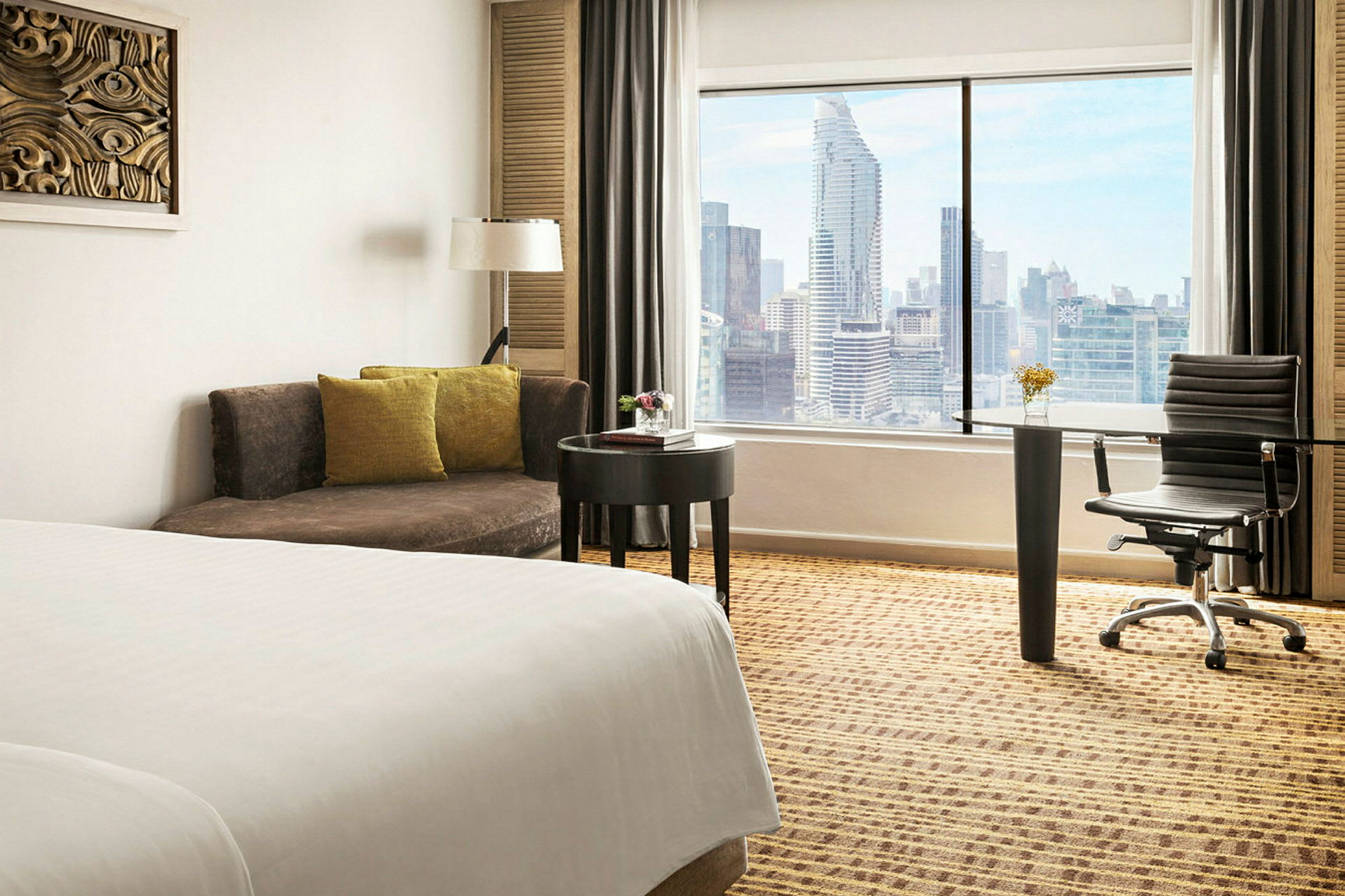 Bild av ett hotellrum med en säng, en soffa, ett skrivbord och en stol. Ett stort fönster visar en vy av en stad. (AI-genererad text)