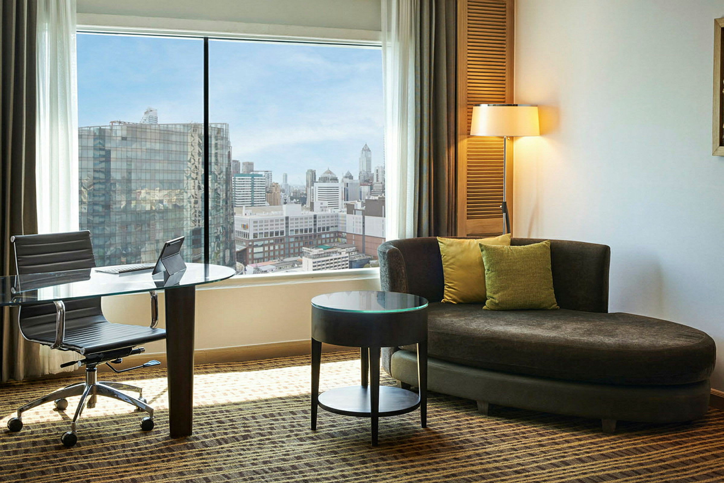 Ett hotellrum med en stor fönstervy över en stad. Det finns en glasbord och stol vid fönstret, samt en brun soffa med kuddar och ett runt sidobord. (AI-genererad text)