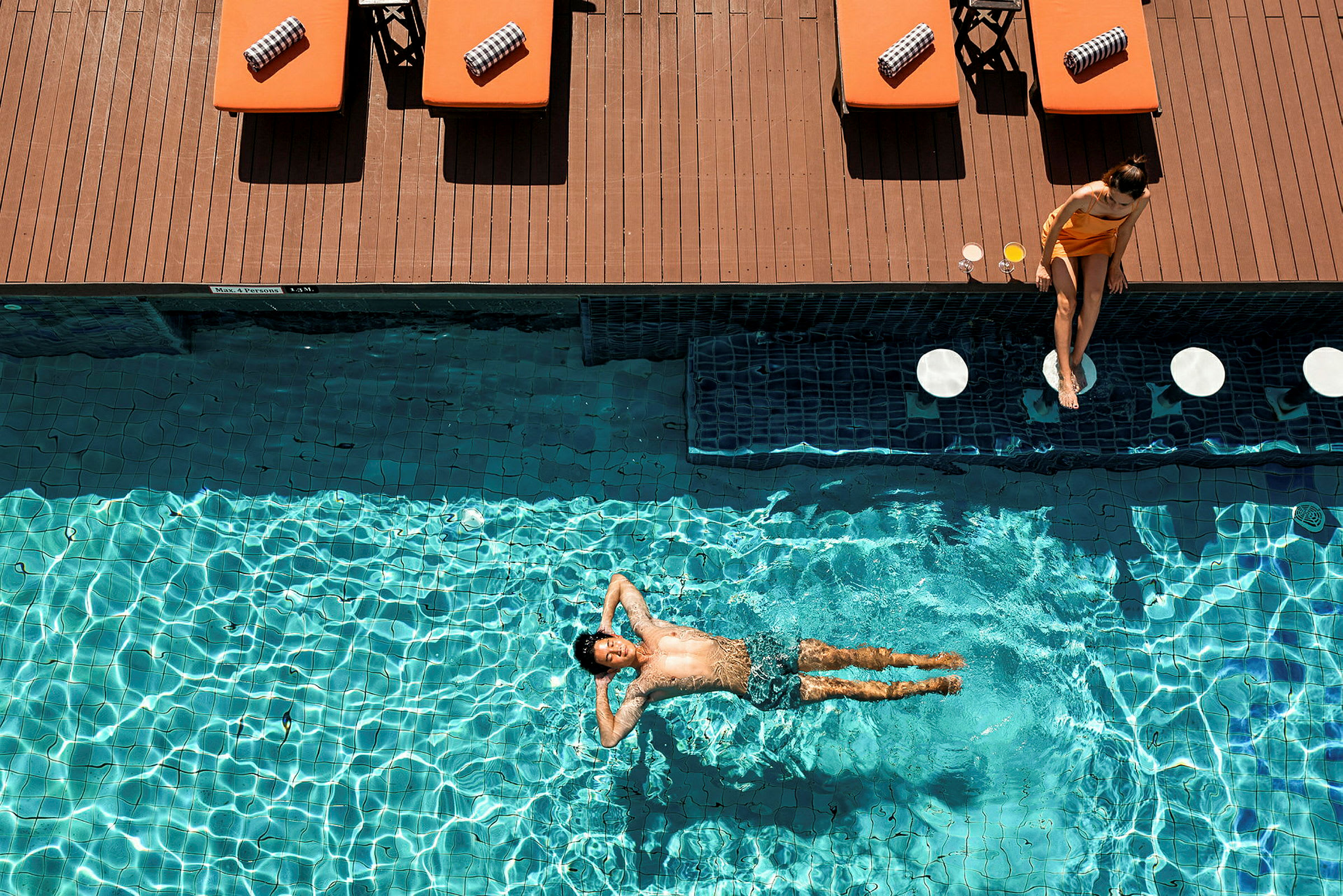 En person ligger och flyter i en pool medan solstolar står längs kanten. (AI-genererad text)