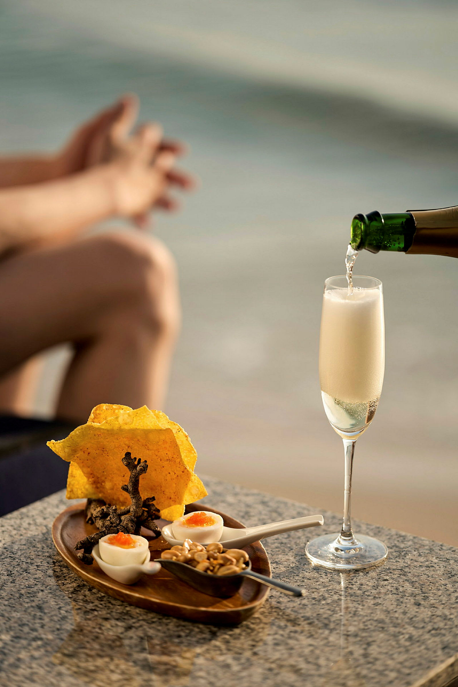 Ett glas champagne hälls upp på ett bord med en liten servering av snacks framför en suddig bakgrund av strand och hav. (AI-genererad text)