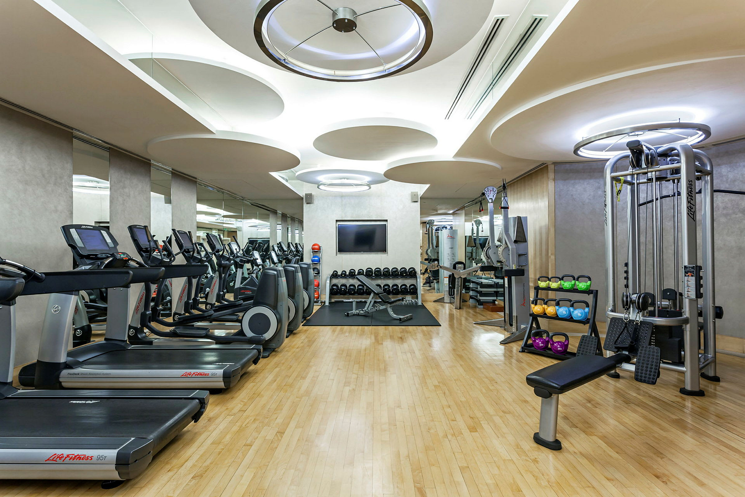 Ett gym med träningsutrustning som löpband, vikter, och träningsmaskiner, omgiven av speglar och med parkettgolv. (AI-genererad text)