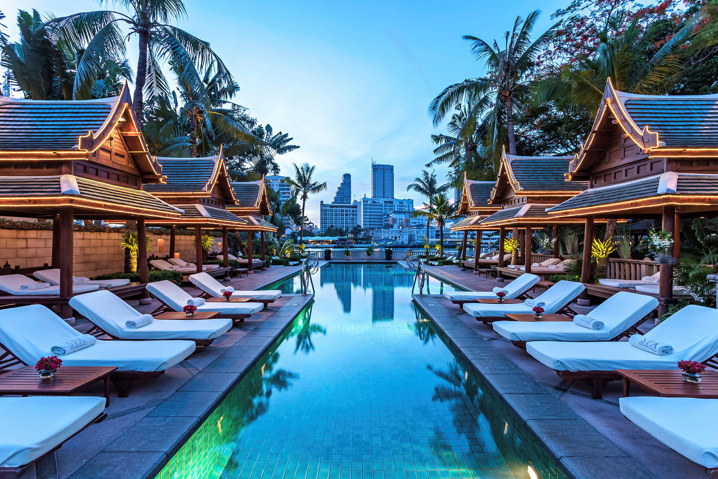 En pool med solstolar på båda sidor, omgiven av paviljonger med thailändsk arkitektur och palmer, kvällsbelysning. (AI-genererad text)