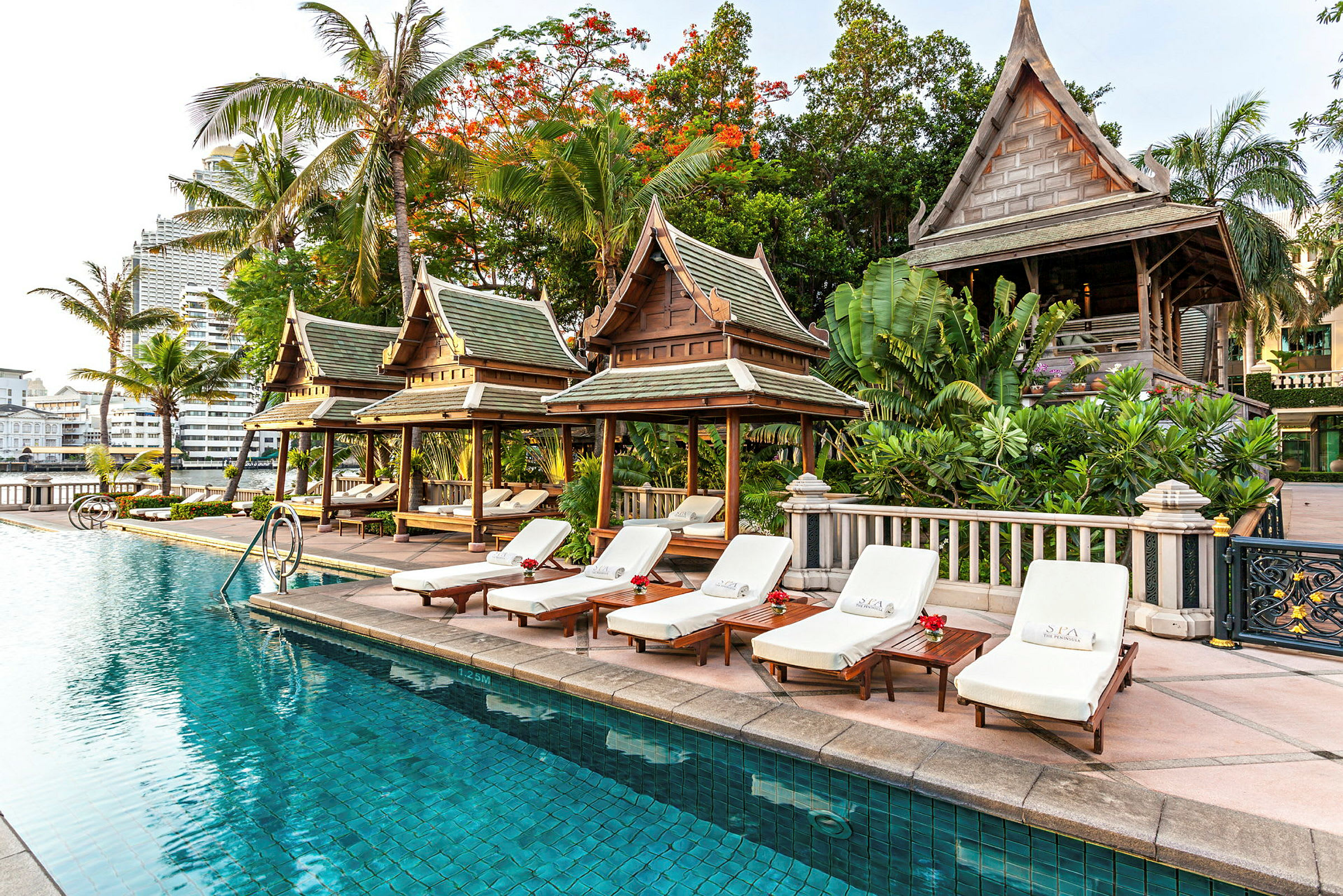 En pool omgiven av solstolar och traditionella thailändska träpaviljonger, omringad av grönska och tropiska träd. (AI-genererad text)