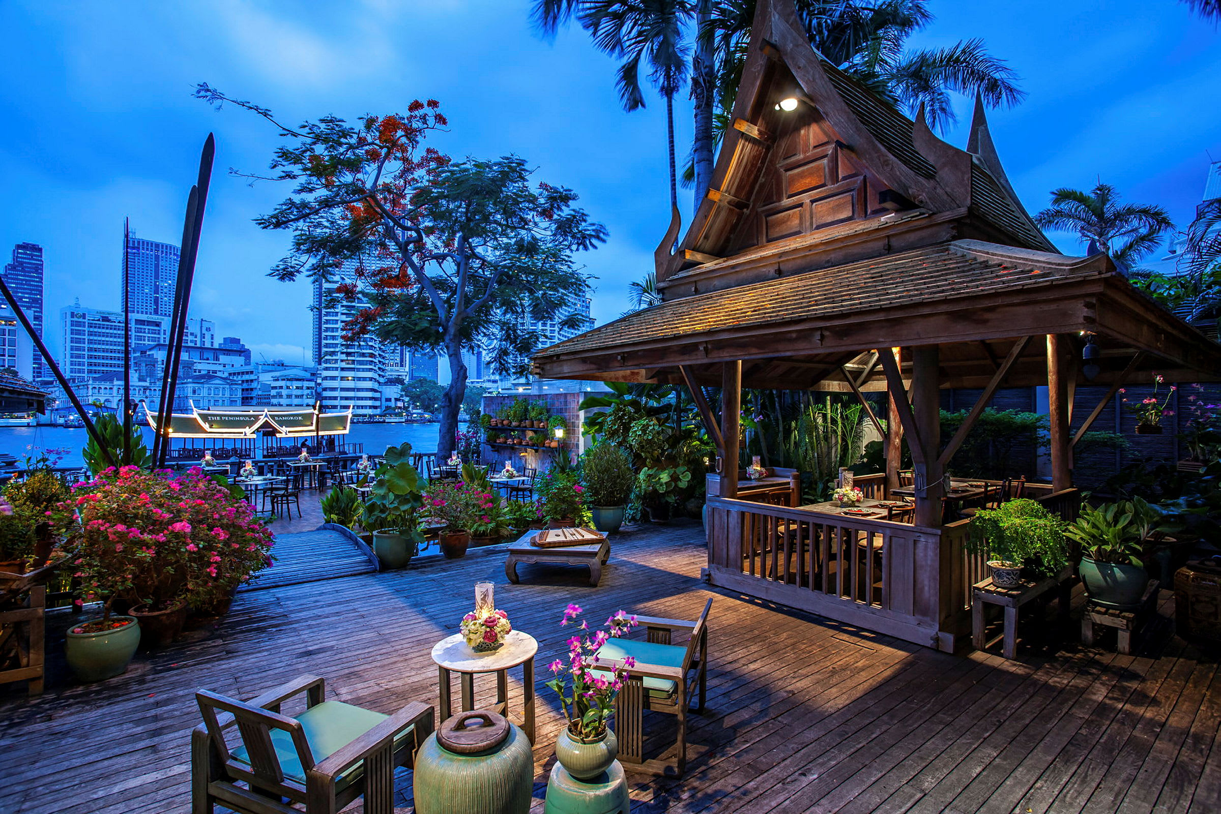 Ett upplyst trähus med traditionell thailändsk design står på en terrass omgiven av blommor och växter. I bakgrunden syns en flod och stadens skyline vid skymningen. (AI-genererad text)