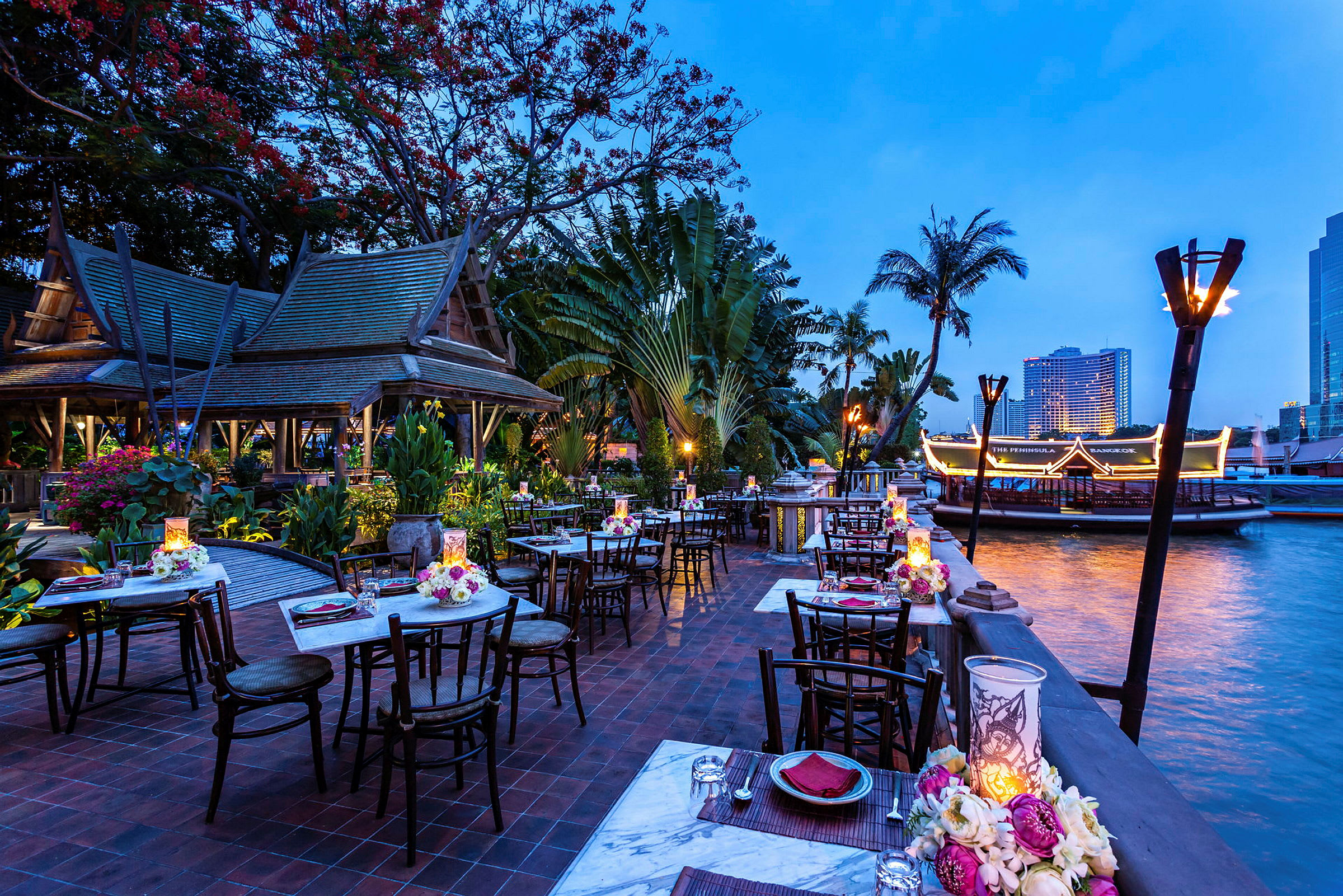 En utomhusrestaurang med uppdukade bord vid en flod. Restaurangen har traditionella thailändska inslag och är omgiven av frodig vegetation. (AI-genererad text)