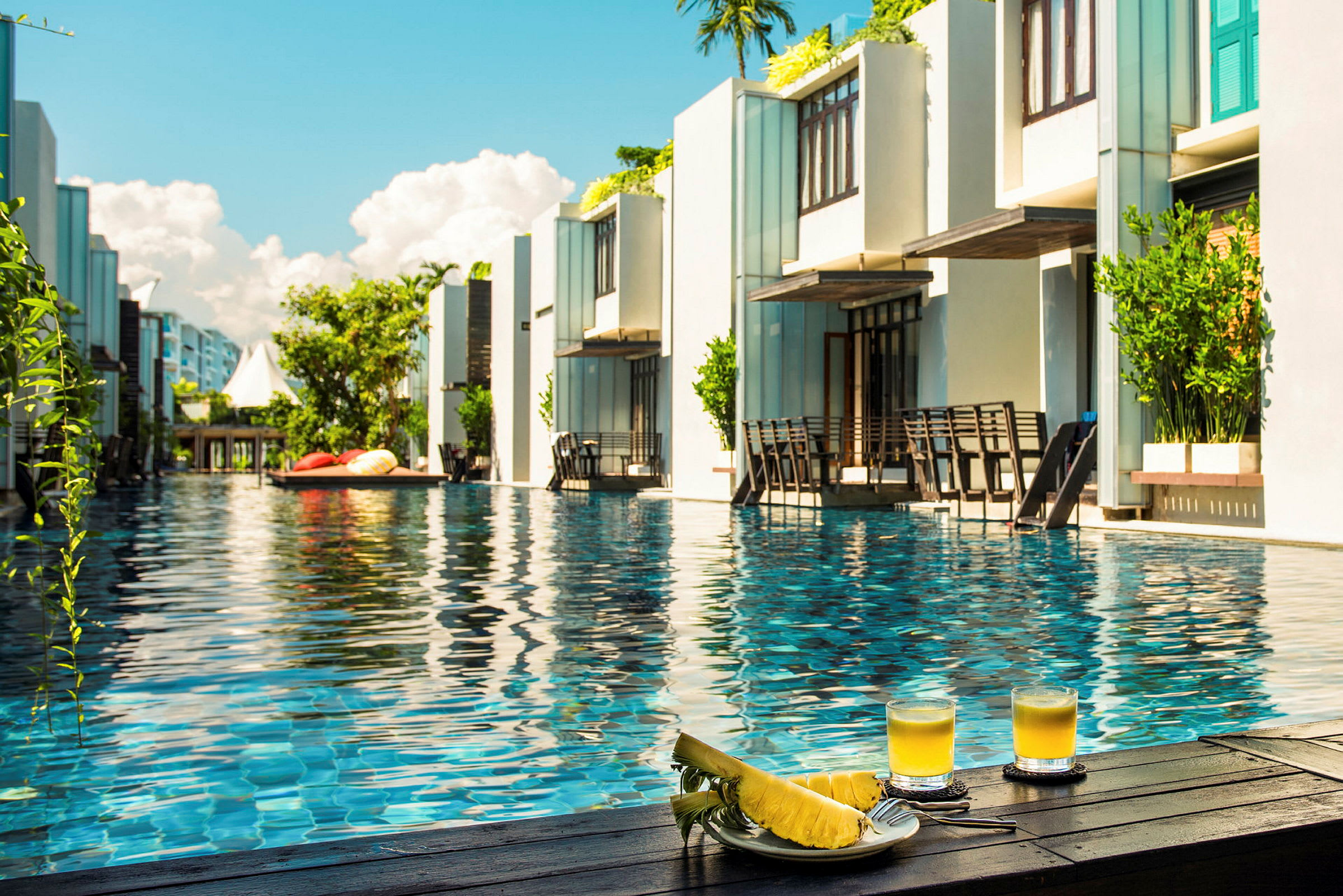 Resortpool omgiven av moderna byggnader med två glas juice och skivat ananas i förgrunden. (AI-genererad text)