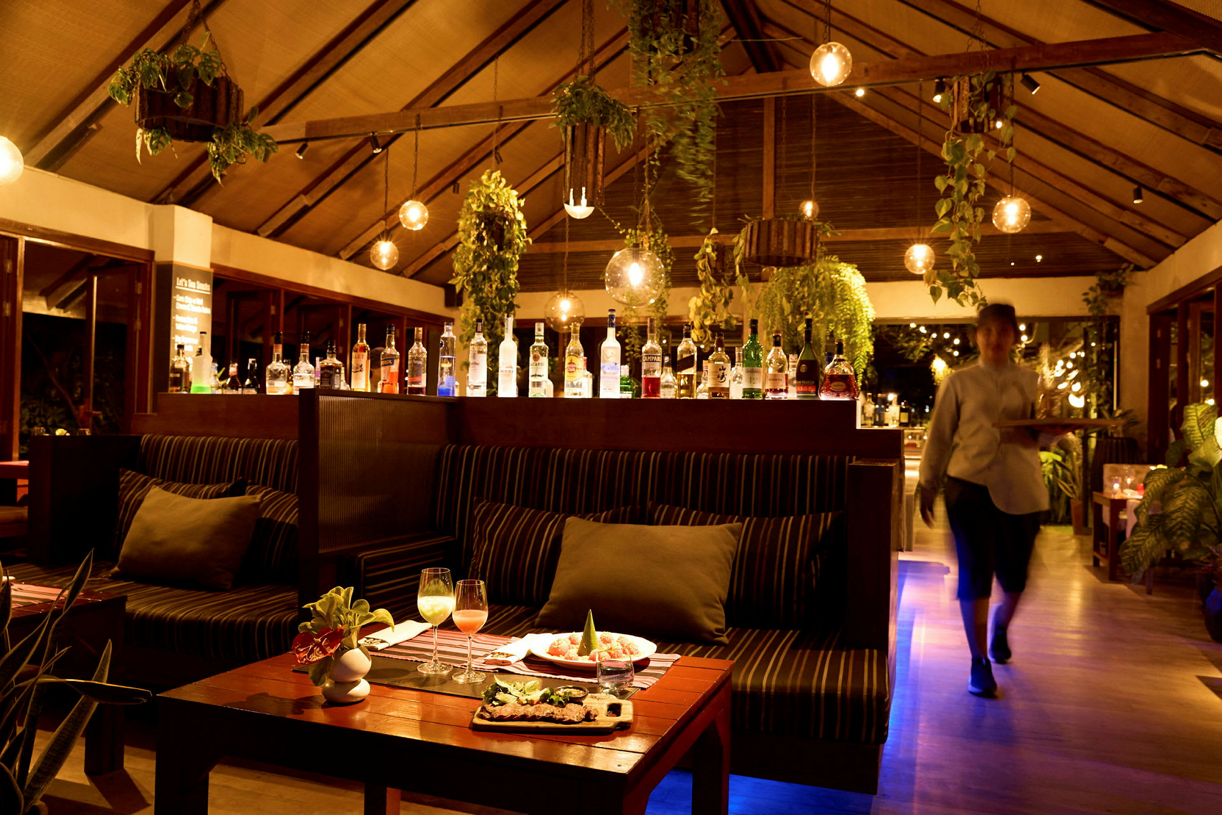 Interiör av en restaurang med sittplatser, hängande växter och en bar som visar upp olika spritflaskor. (AI-genererad text)