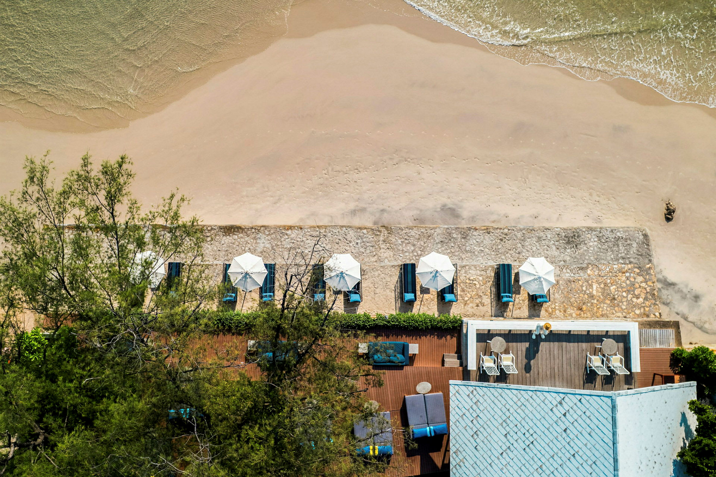 Vy från ovan av en sandstrand med parasoller och solstolar samt en närliggande byggnad med terass. (AI-genererad text)