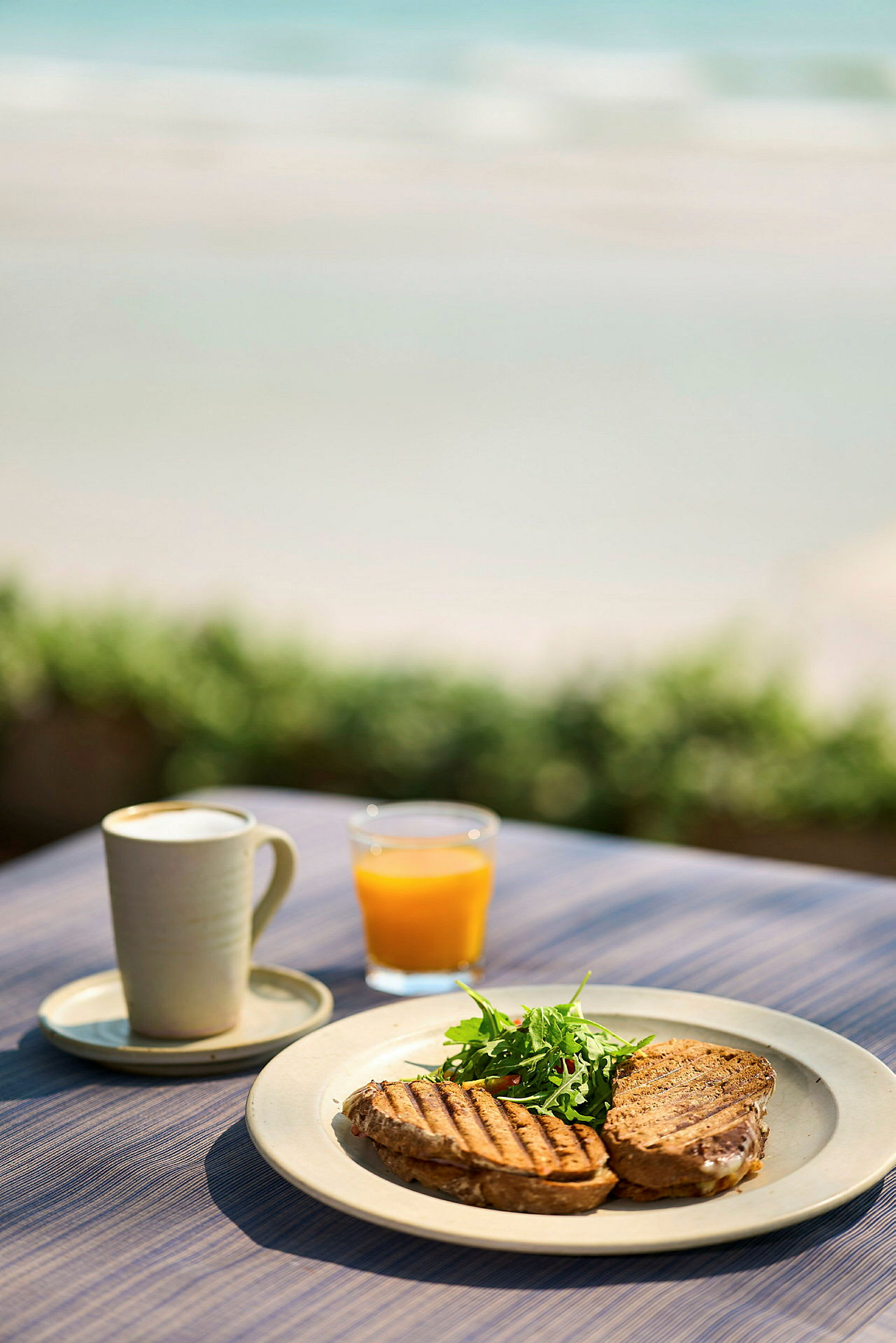En tallrik med grillade smörgåsar och sallad, ett glas apelsinjuice och en kopp kaffe står på ett bord vid stranden. (AI-genererad text)