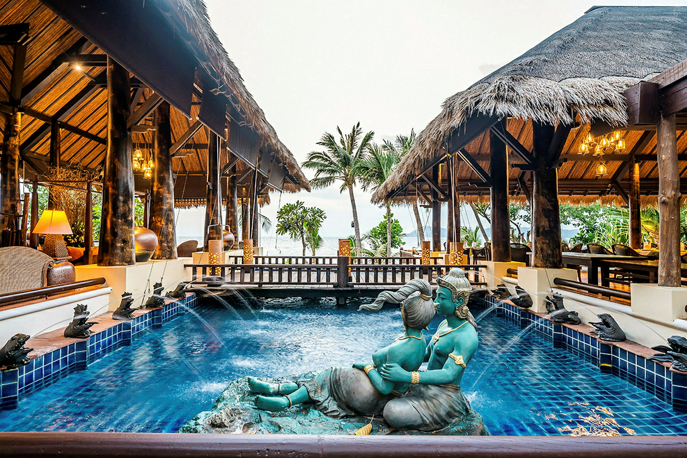 En pool med en skulptur i förgrunden omgiven av traditionellt byggda paviljonger med halmtak, palmer och utsikt mot havet. (AI-genererad text)