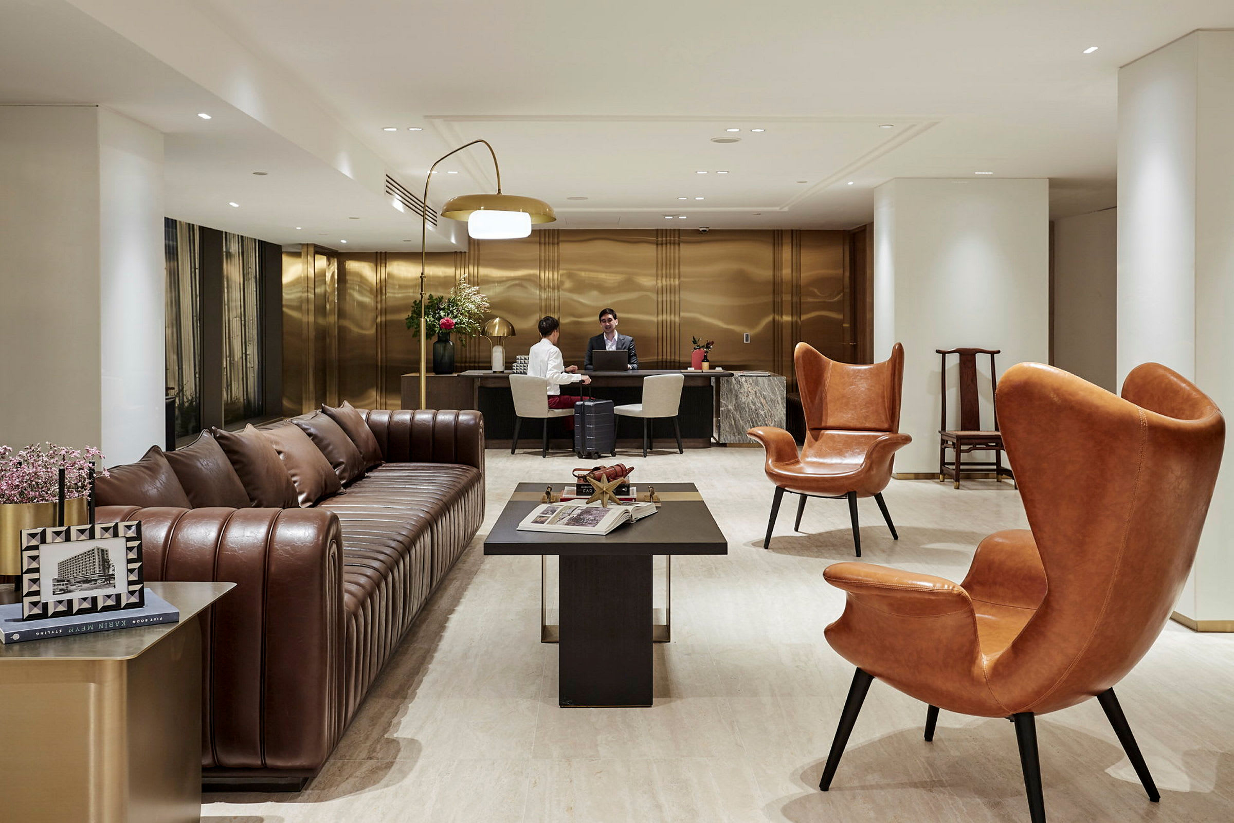 Ett hotellobbyområde med en brun lädersoffa, orange läderfåtöljer och ett mörkt soffbord framför. I bakgrunden finns en receptionsdisk där två personer samtalar. (AI-genererad text)