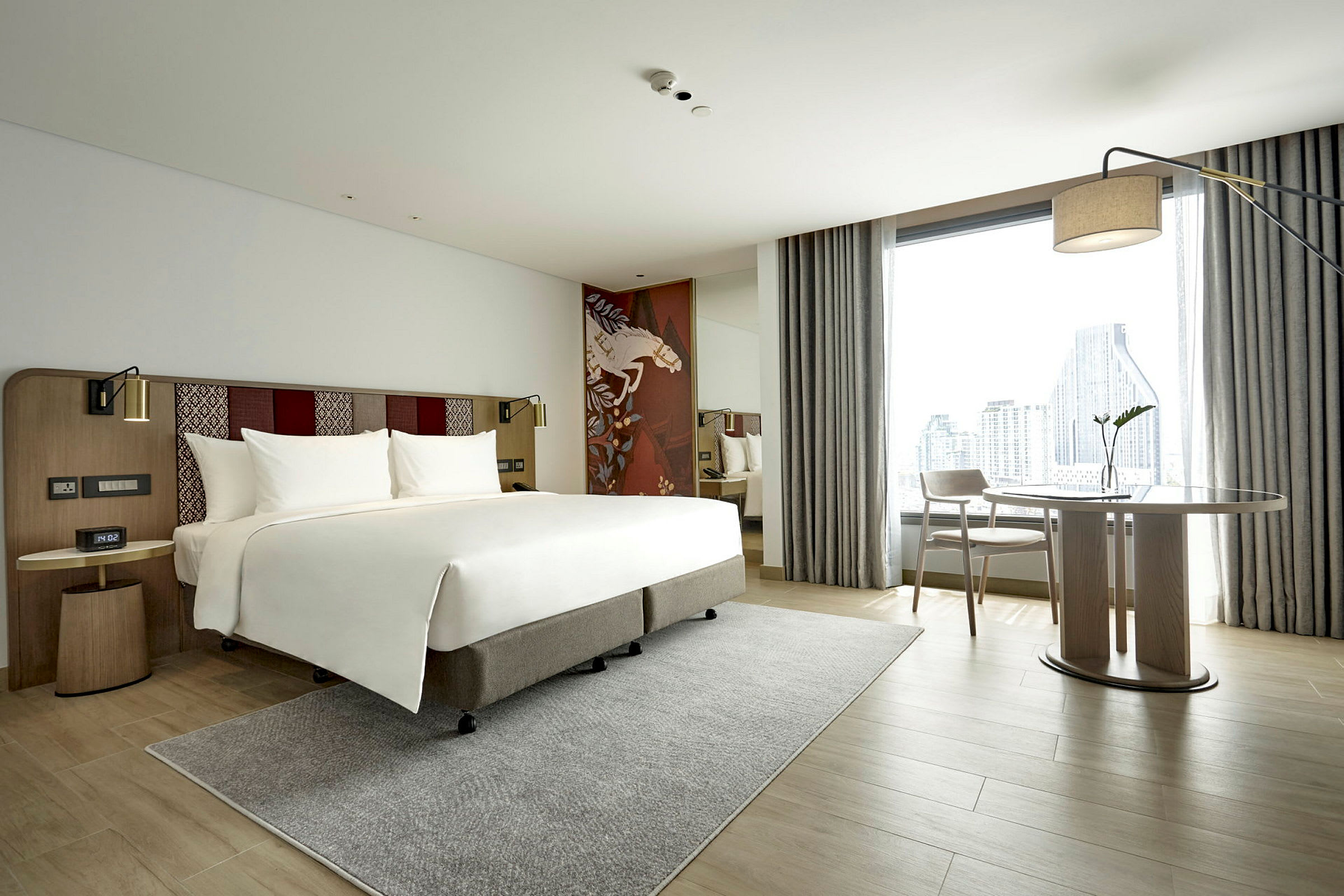 Modernt hotellrum med en dubbelsäng, skrivbord och utsikt över en stad genom ett stort fönster. (AI-genererad text)