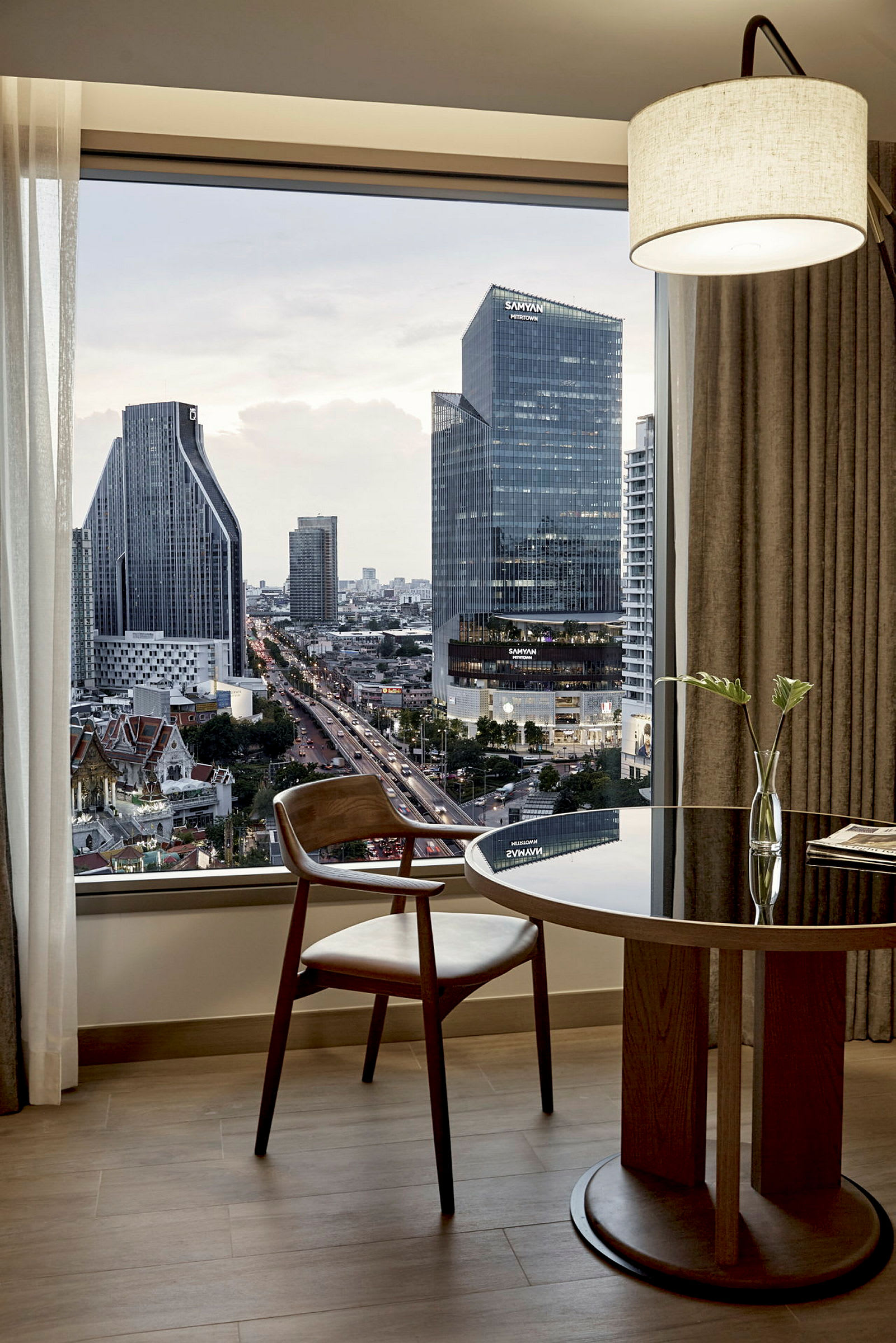 Ett modernt hotellrum med utsikt över en stad med höga byggnader i kvällsljus. Ett fönster visar utsikten, och framför det står ett träbord med en stol samt en enkel blomstervas. (AI-genererad text)
