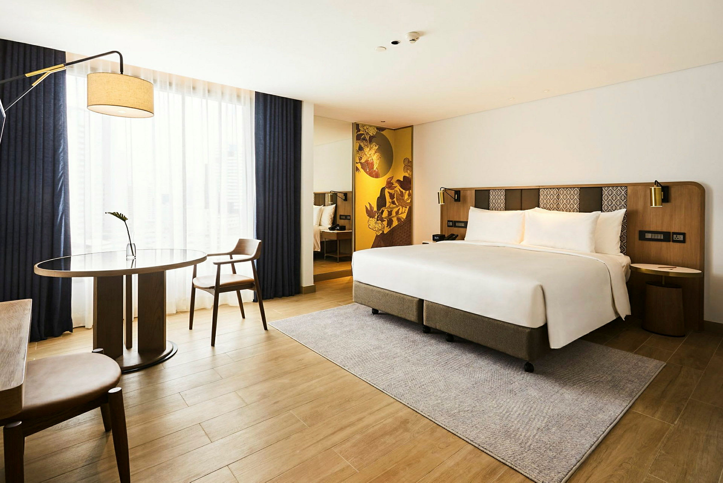 Ett modernt hotellrum med en dubbelsäng, ett bord och två stolar samt en stor fönsteryta med gardiner. (AI-genererad text)