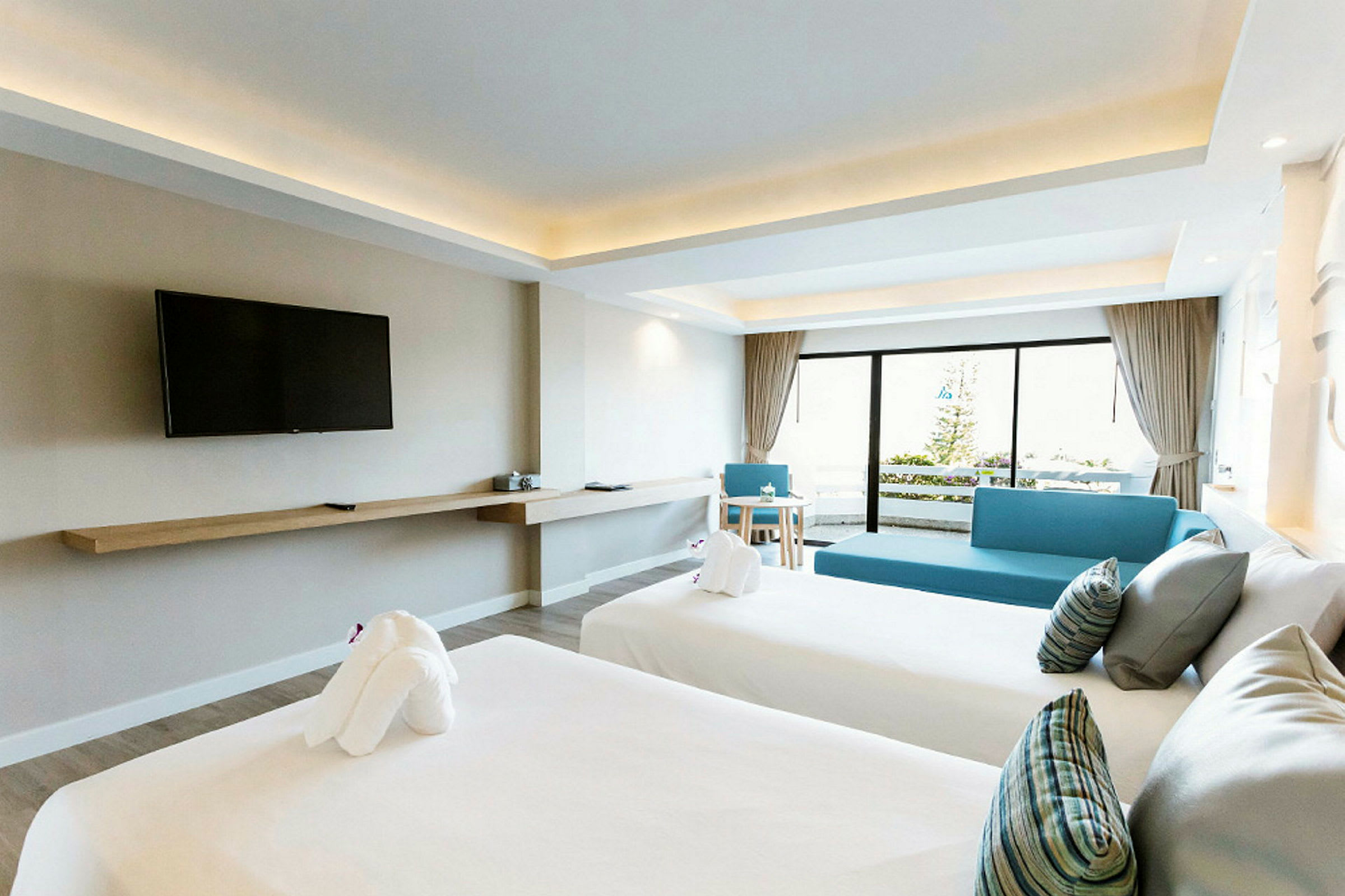 Interiör av ett hotellrum med två bäddade sängar, en väggmonterad TV och ett område med sittmöbler med utsikt mot en balkong. (AI-genererad text)