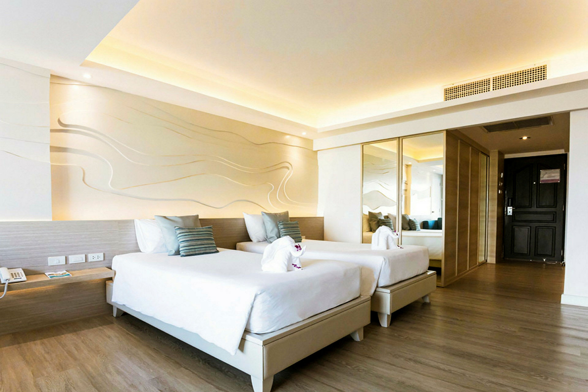 Ett hotellrum med två enkelsängar, vit inredning och trädetaljer. Väggarna har dekorativa mönster och det finns en spegel samt en svart dörr. (AI-genererad text)
