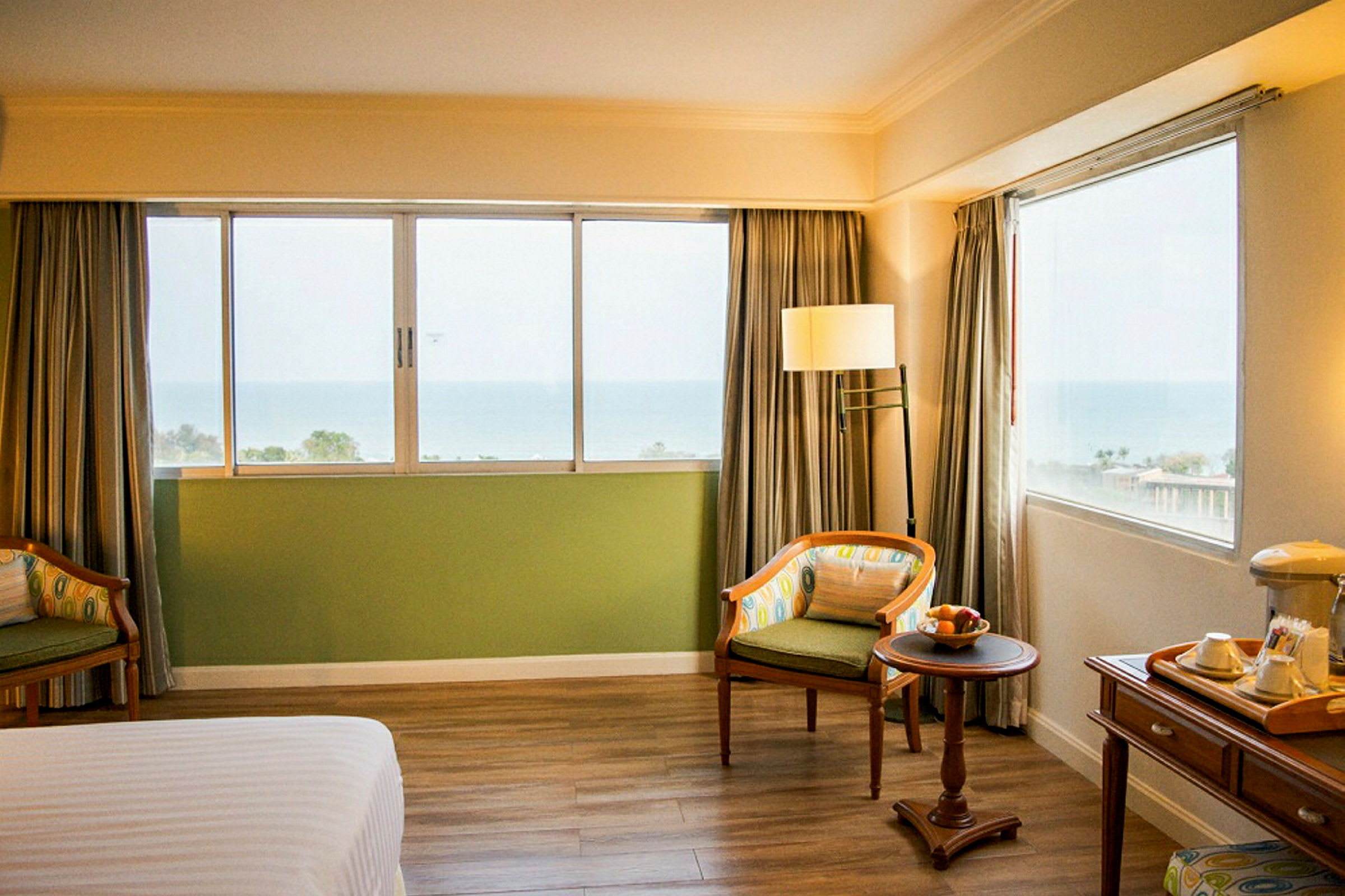 Ett hotellrum med två fåtöljer, ett litet bord med frukt, ett fönster med havsutsikt och ett skrivbord med tebricka. (AI-genererad text)