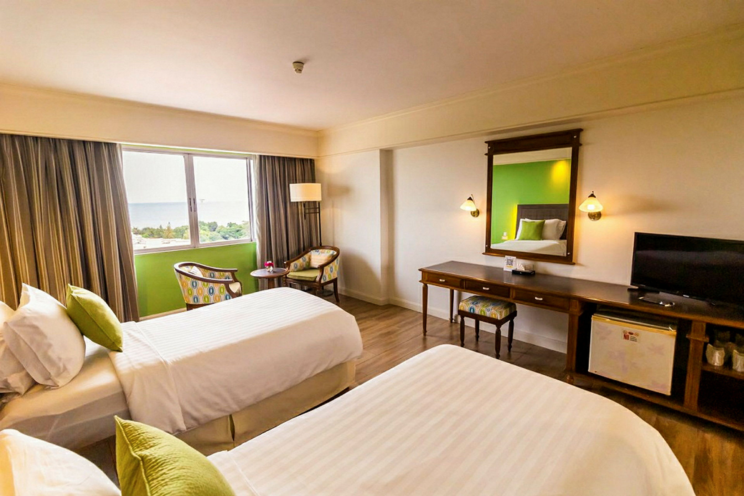 Ett hotellrum med två enkelsängar, färgglada kuddar, ett skrivbord med spegel och en TV samt en sittgrupp vid ett fönster. (AI-genererad text)